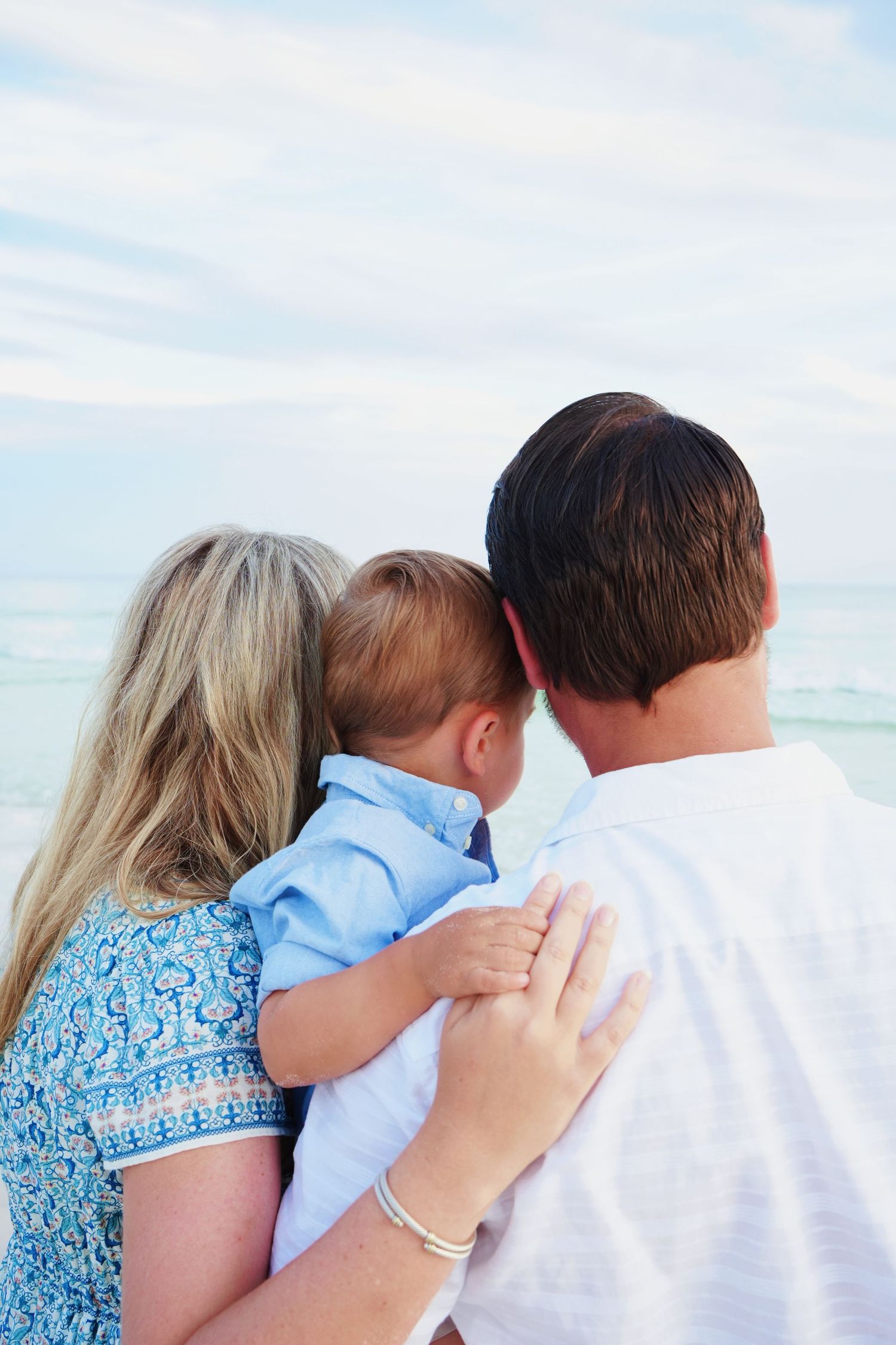 A family embraces while standing together on a sunny beach, wearing coordinating blue and white outfits.