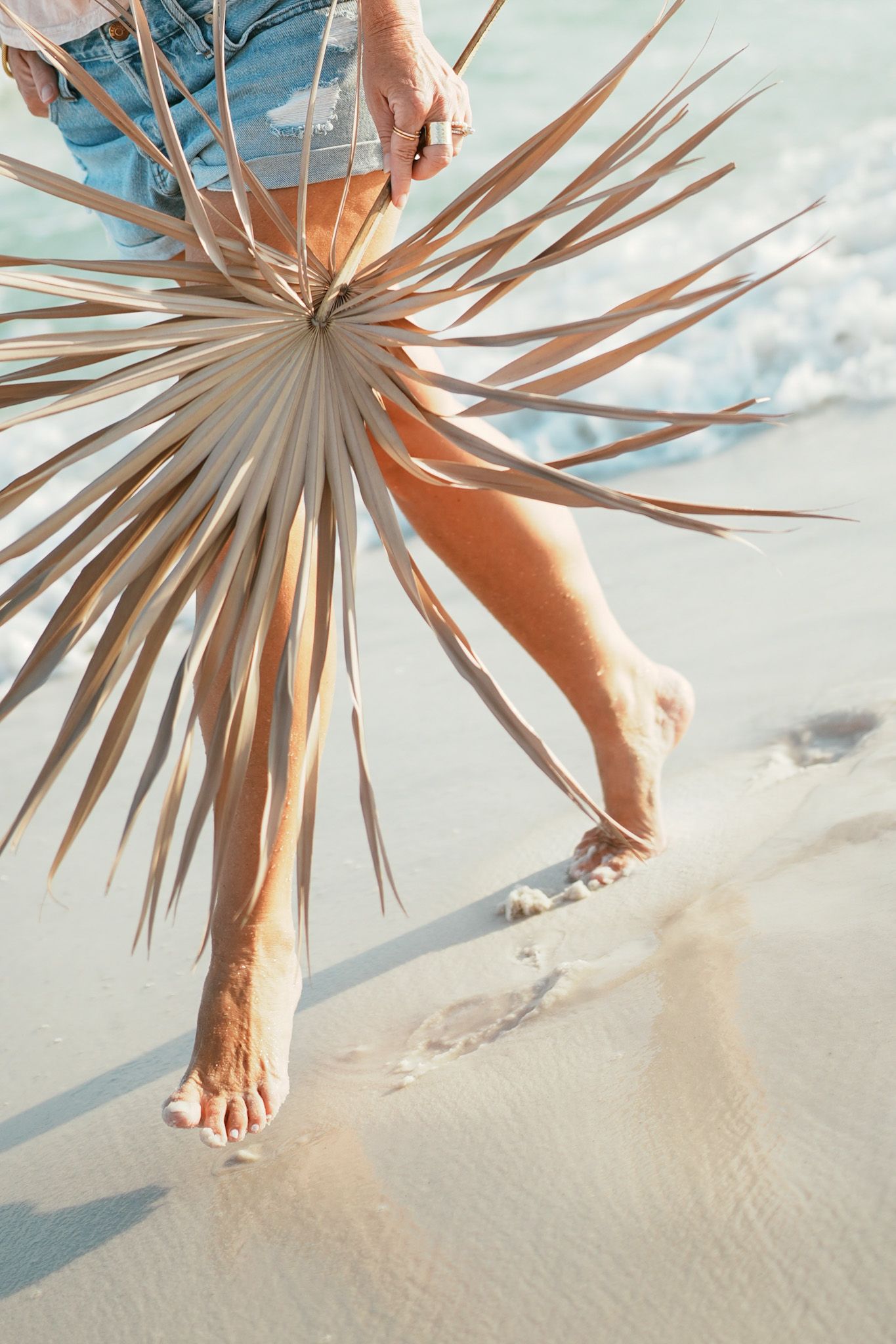 Bare feet walking on sandy beach with dried palm frond casting shadows against cloudy sky.