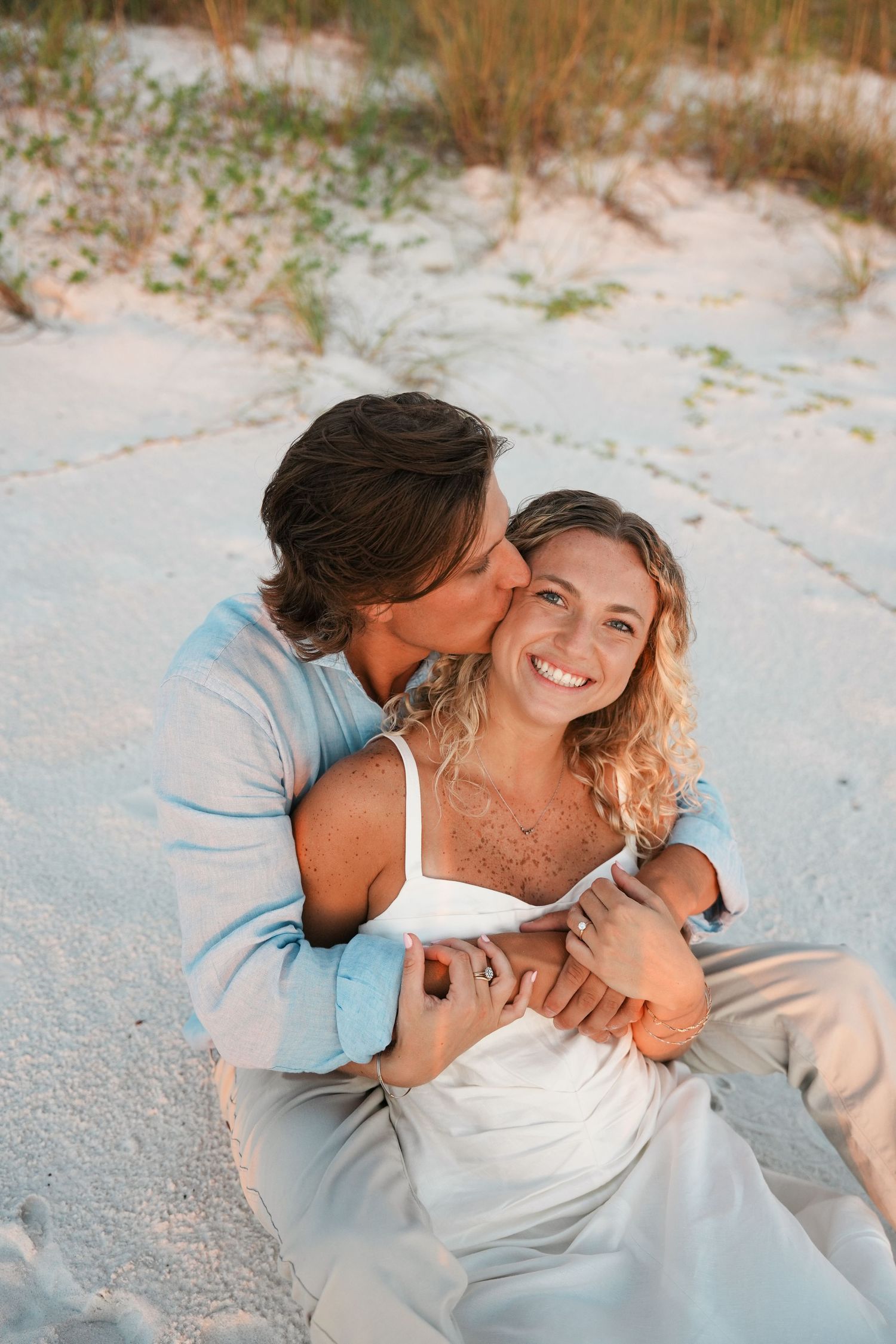 A romantic couple shares tender moments while sitting together on white sandy beach.