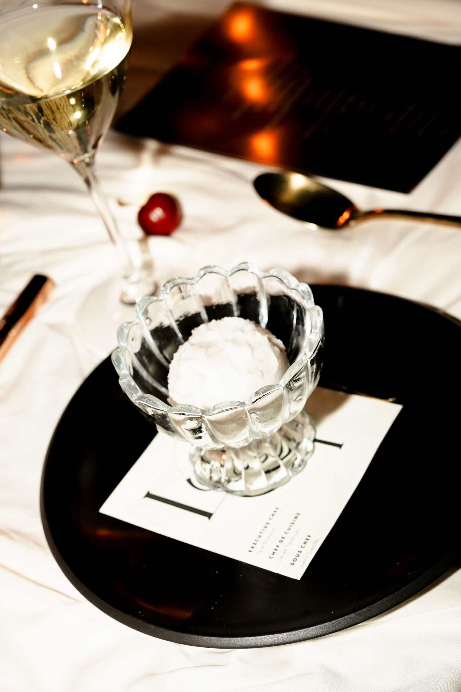 Elegant dessert table setting with ice cream in crystal dish, champagne glass, black plate, and gold cutlery.