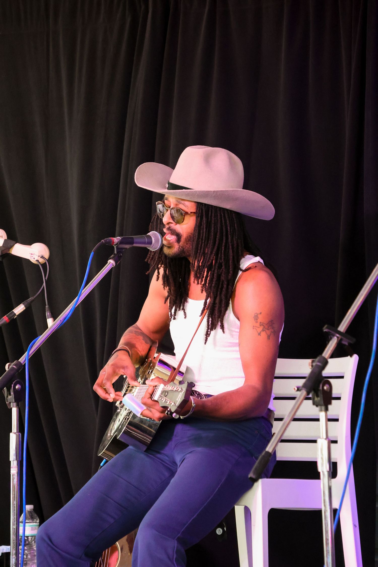 Musician performing on acoustic guitar wearing pink cowboy hat and white tank top on stage.