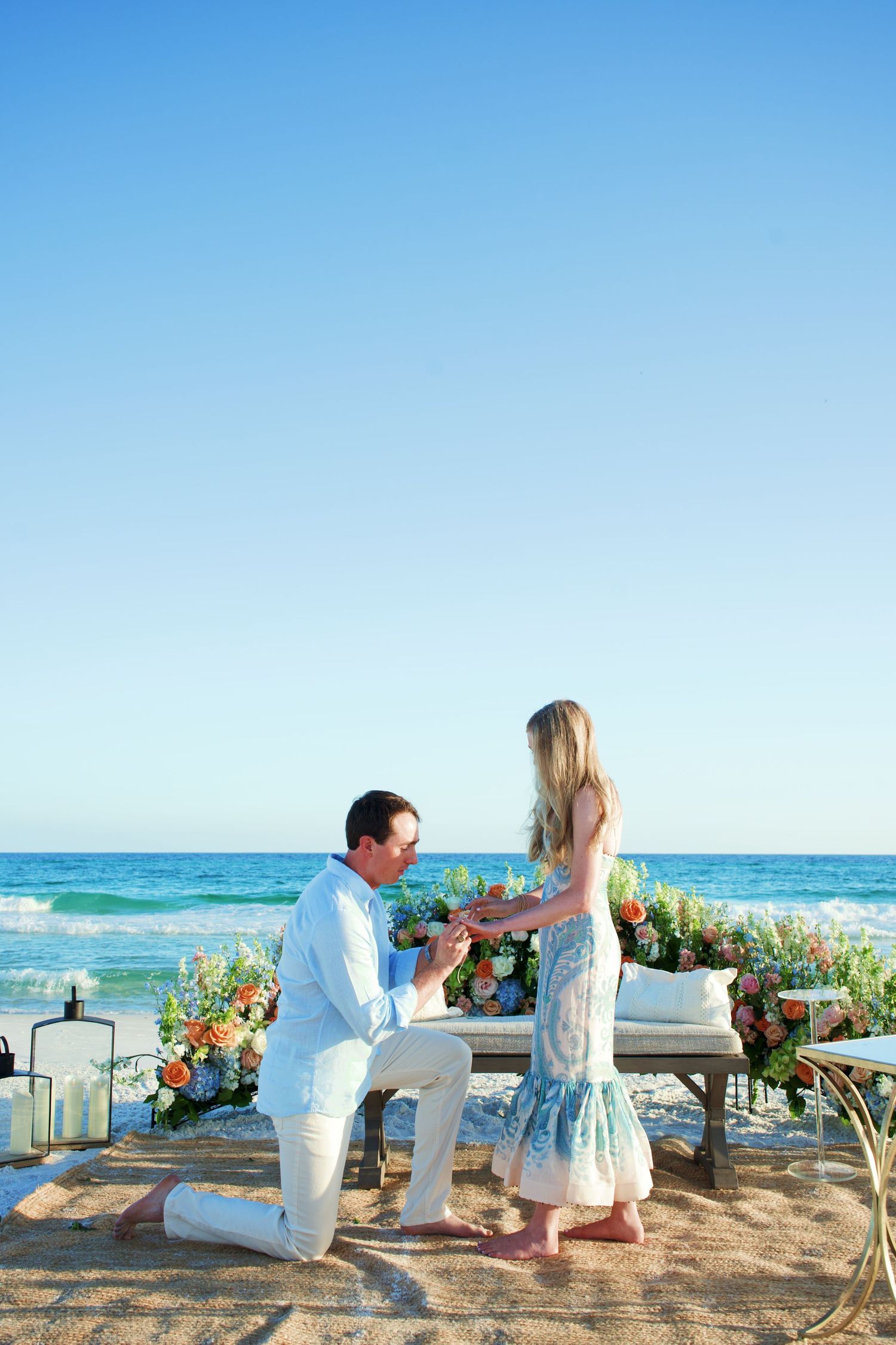 A beach proposal setup with flowers and lanterns by the ocean during a bright sunny day.