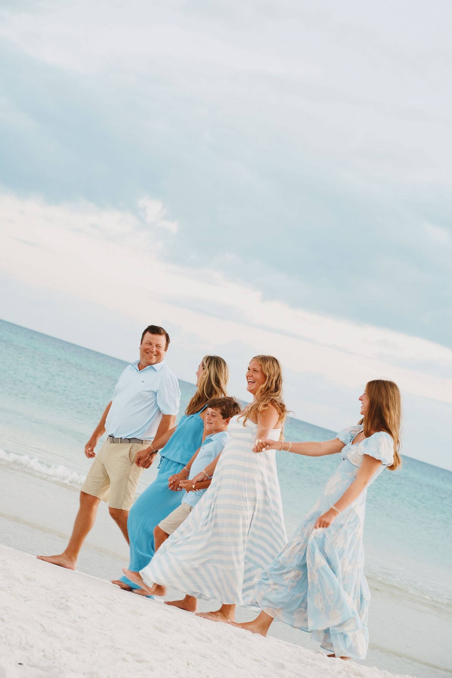 A joyful family group wearing white and blue outfits walks together hand-in-hand along a pristine beach at sunset.