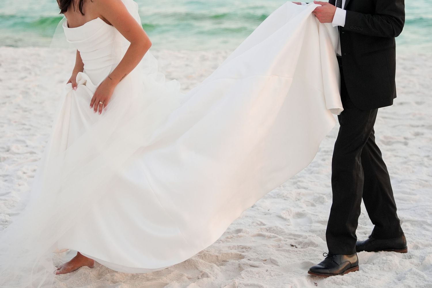 Bride and groom on a sandy beach, with the groom holding the flowing white wedding dress train.