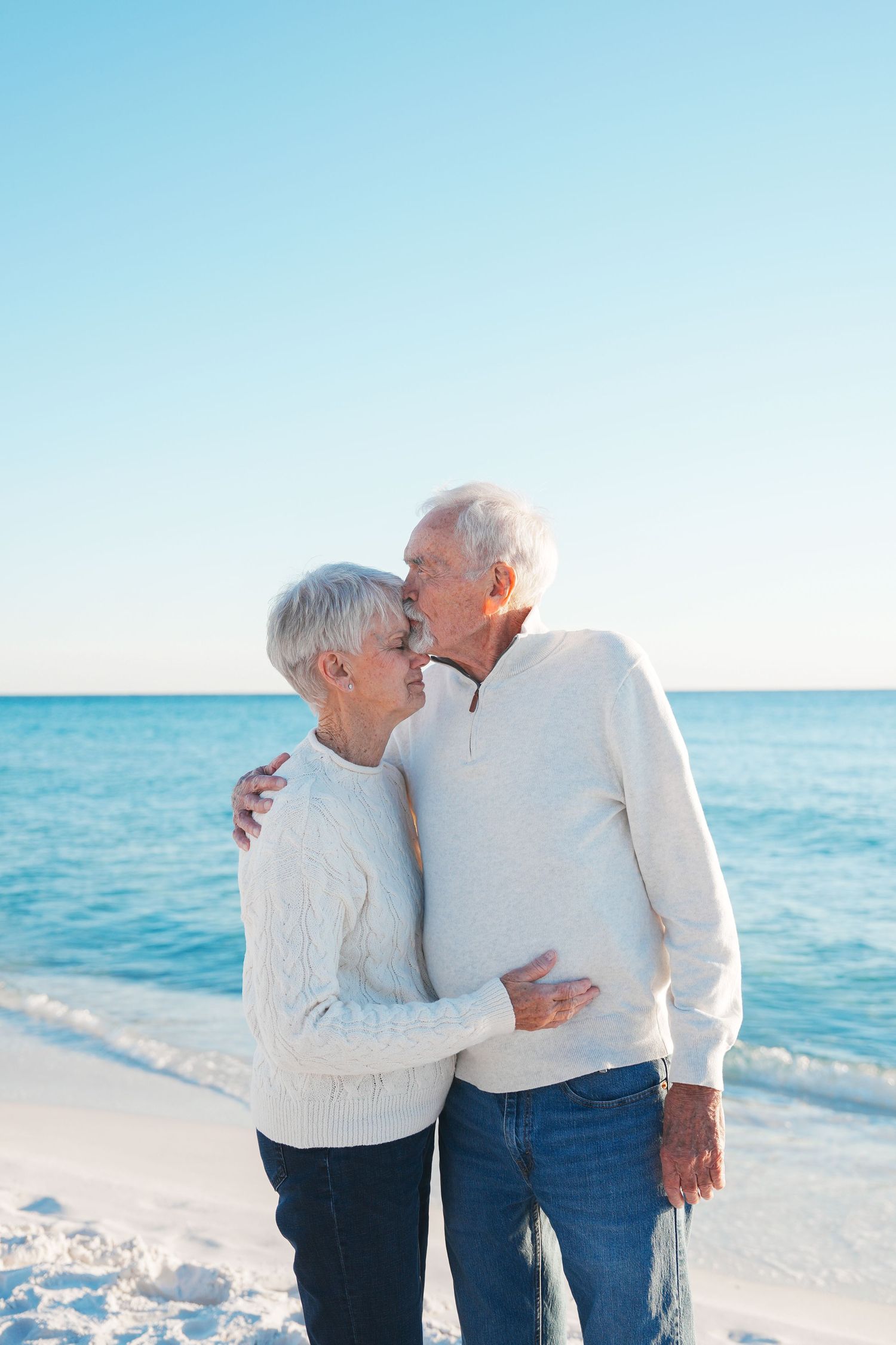 A loving couple in white sweaters embrace while standing on a sunny beach with turquoise ocean waters in the background.