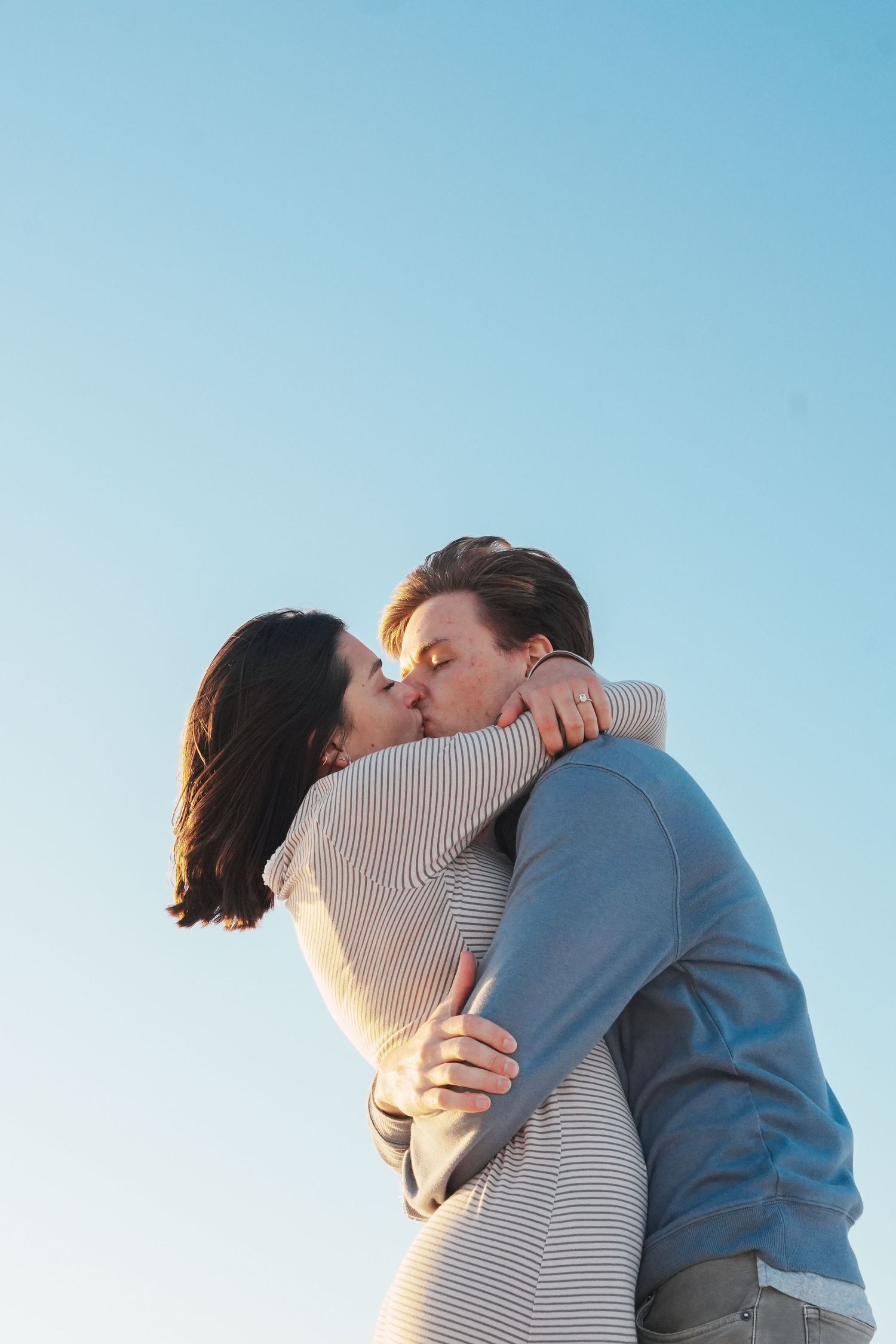 A couple embraces and kisses against a bright blue sky background in a romantic outdoor setting.