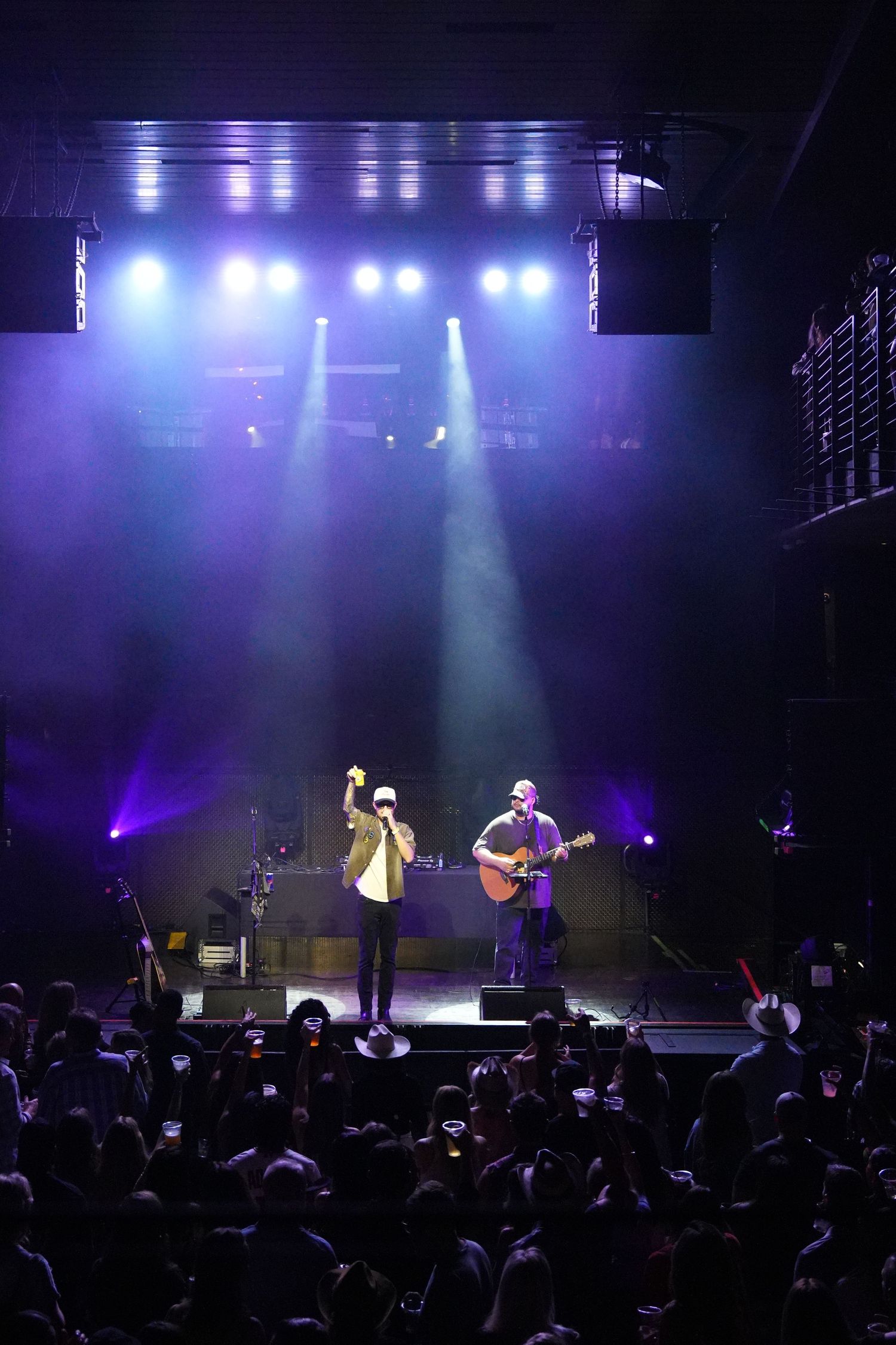 Concert stage illuminated with dramatic purple and blue lighting as performers entertain a crowded audience.