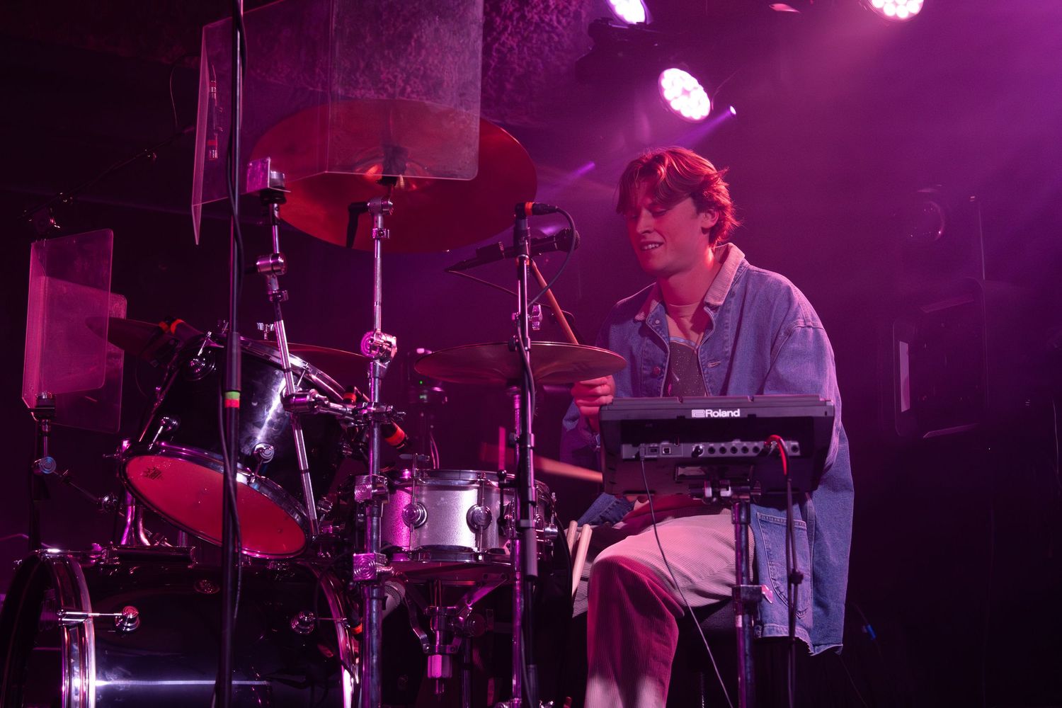 A musician performs on stage with a keyboard setup under purple lighting at a concert venue.
