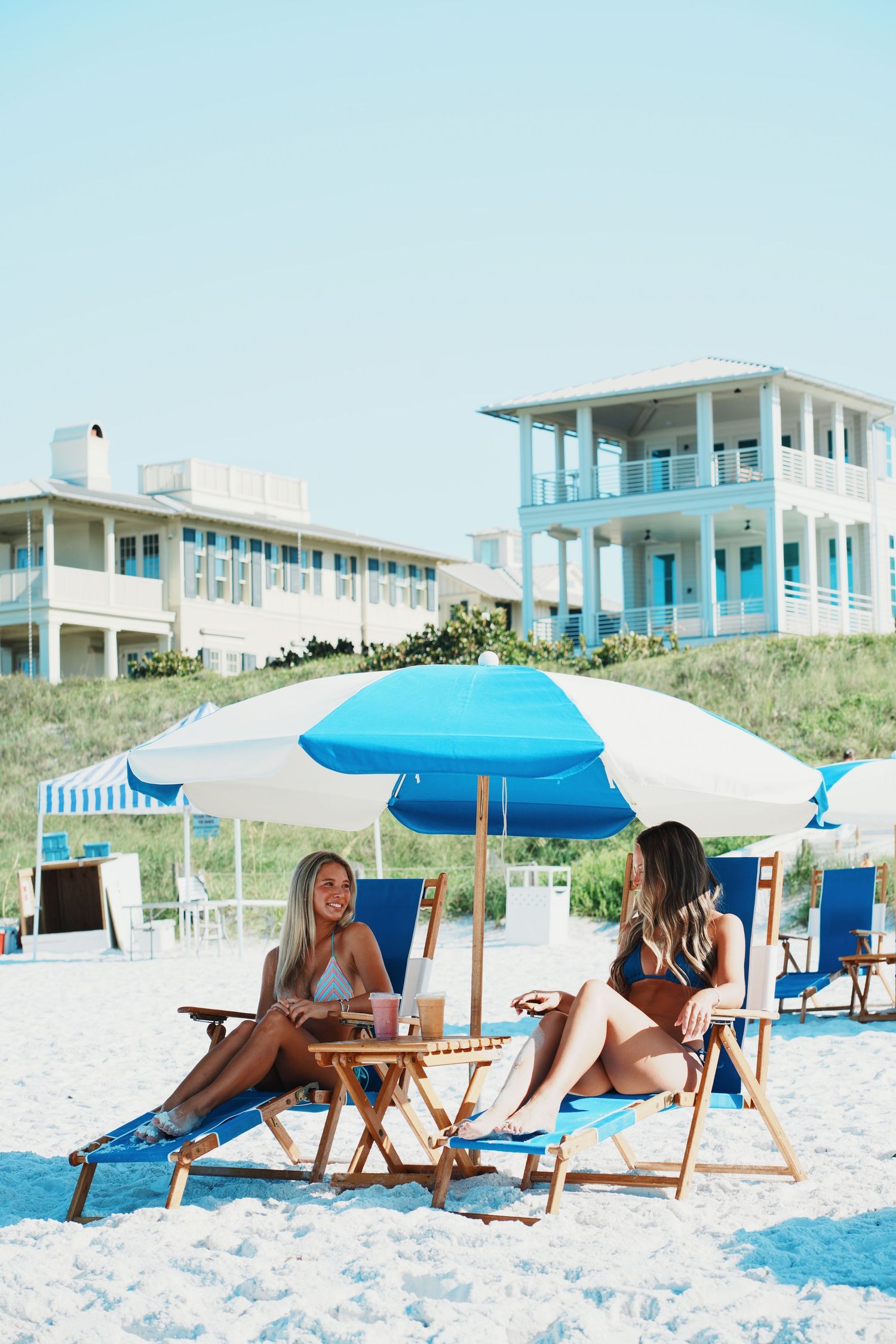 Two beach chairs with umbrella on white sand beach near coastal vacation homes painted in pastel blue tones.