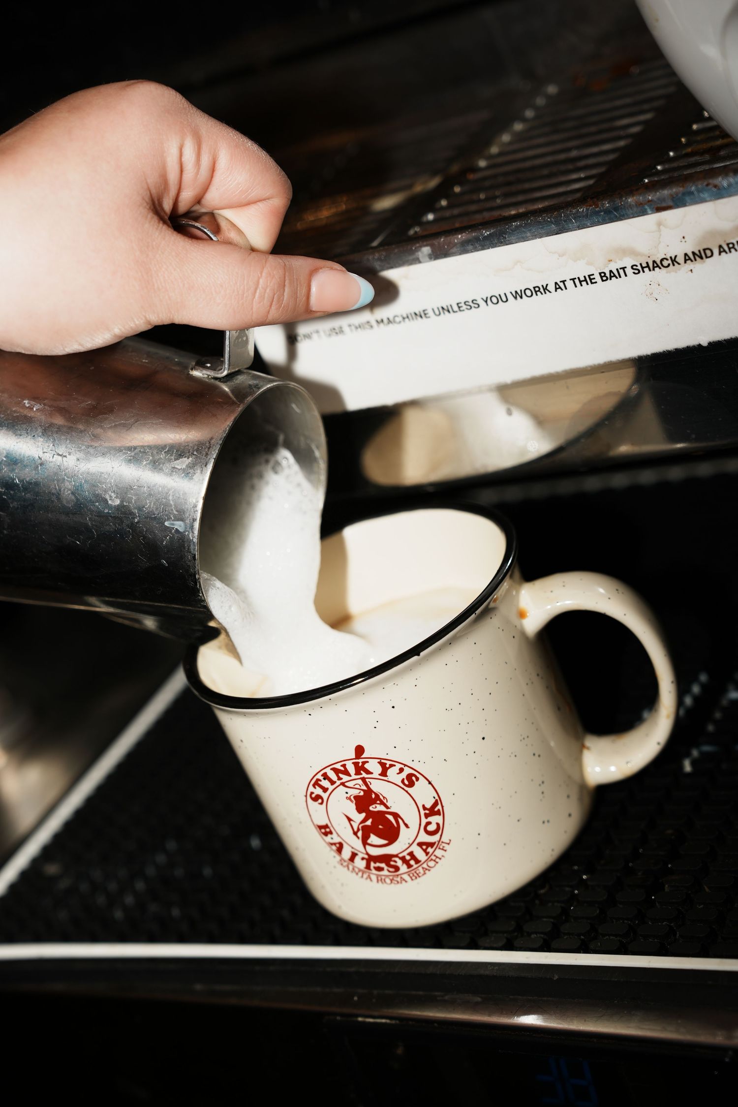 Steamed milk being poured into a vintage white Buckeye's Brewing Co mug on a dark stovetop surface.