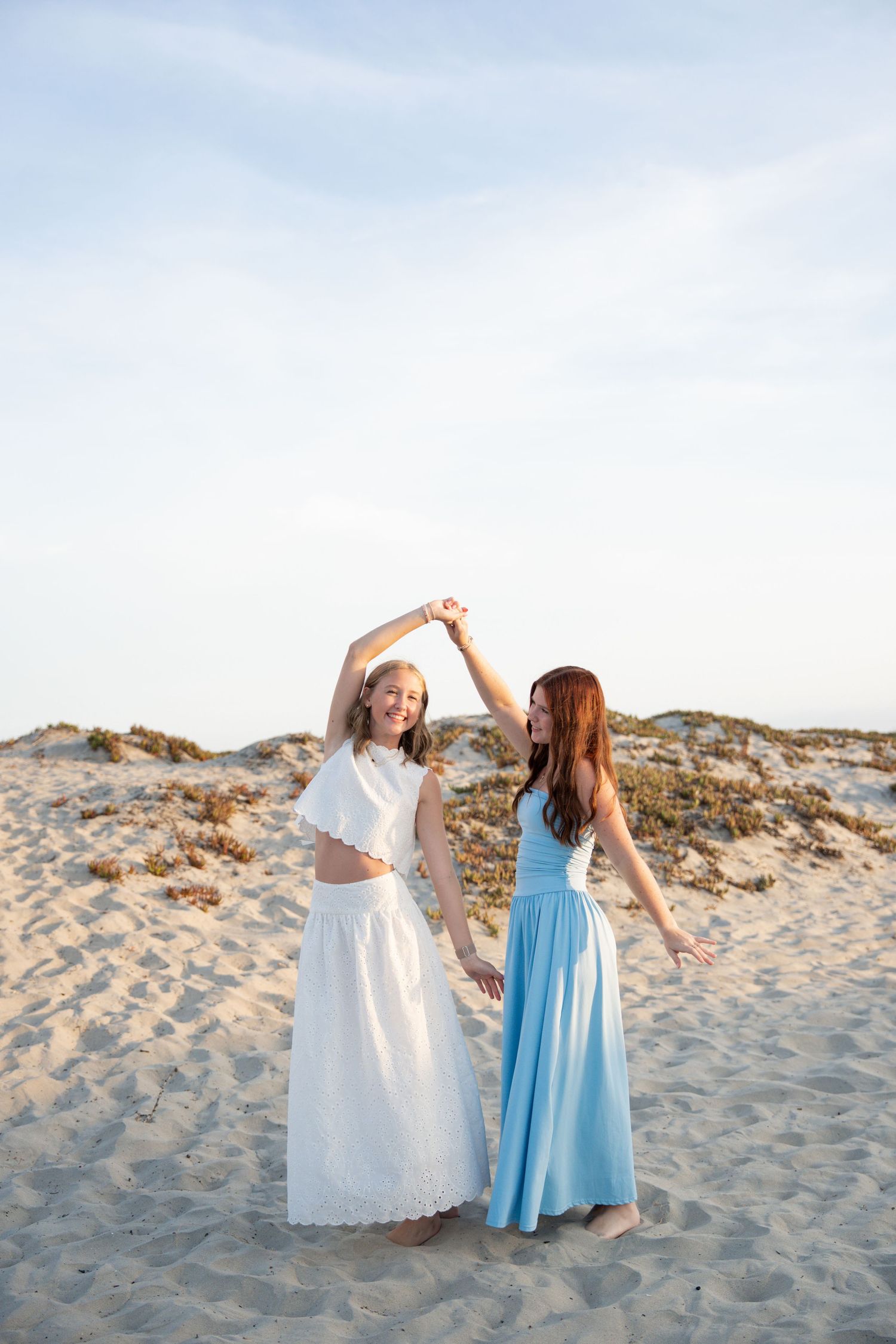Two women in white and blue maxi dresses dancing joyfully on a sandy beach under a clear sky.