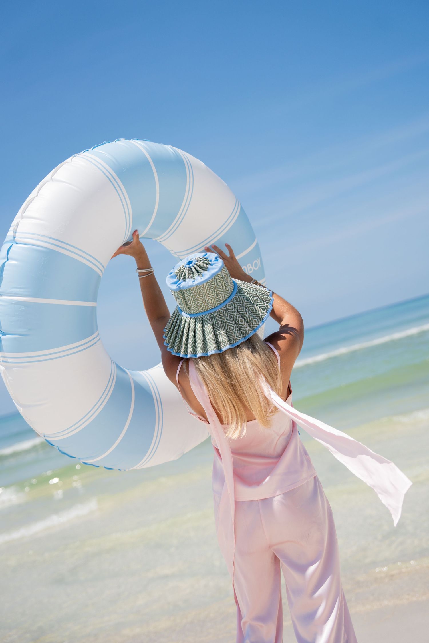A young girl in a pink outfit holds a large striped inflatable ring on a sunny beach.