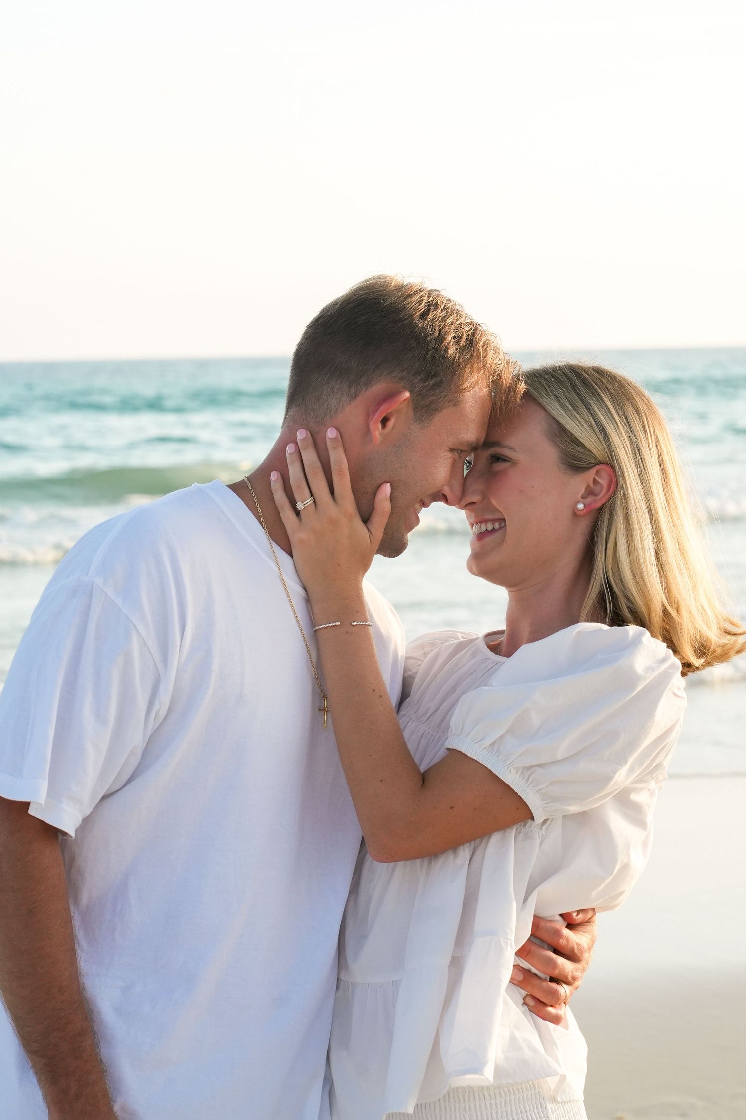 Happy couple in white outfits embracing and smiling on a beach with ocean waves in the background.