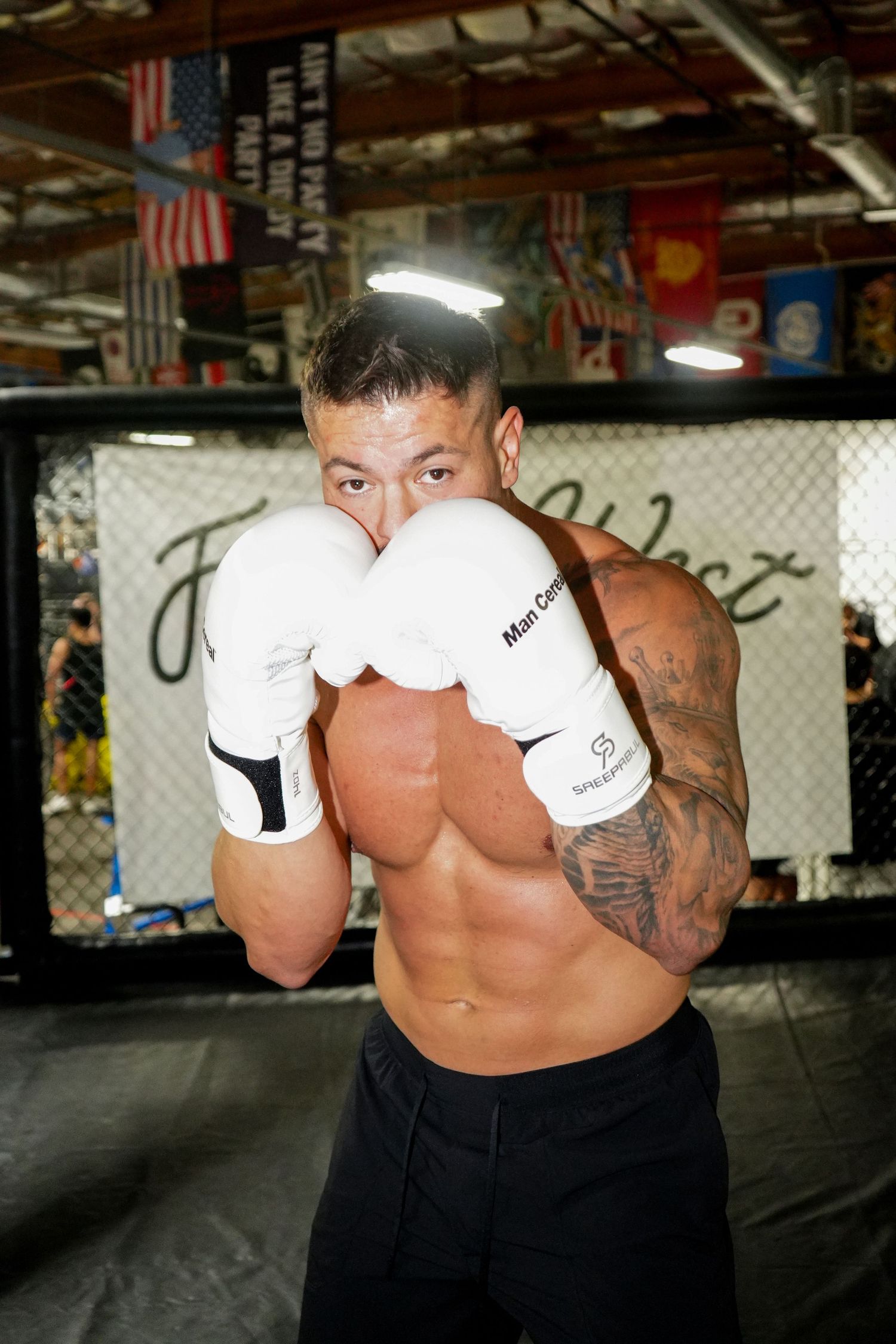 Shirtless boxer in training stance with white gloves and tattoos inside gym with flags hanging from ceiling.