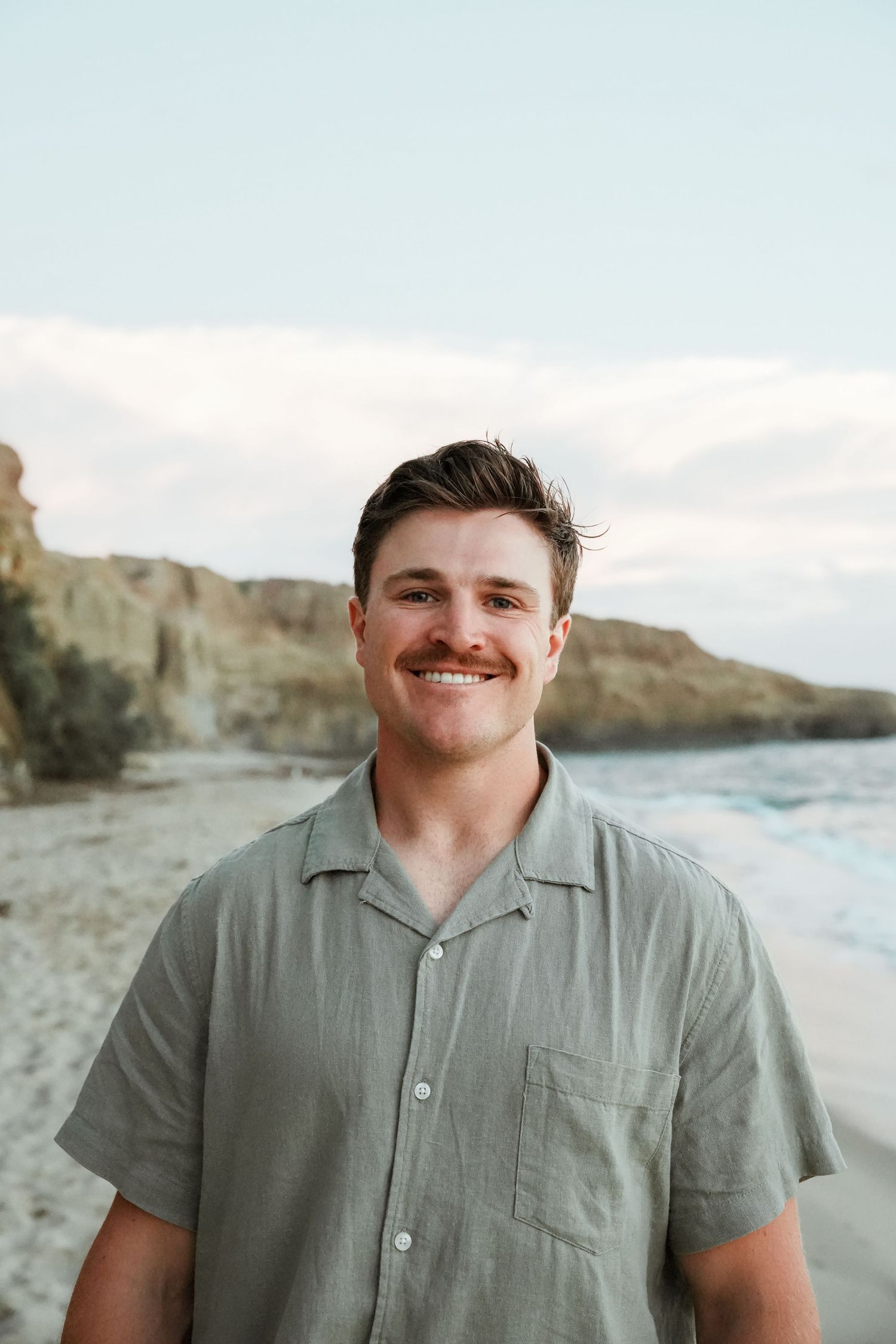 A person in a sage green polo shirt stands on a beach with coastal cliffs in the background at sunset.