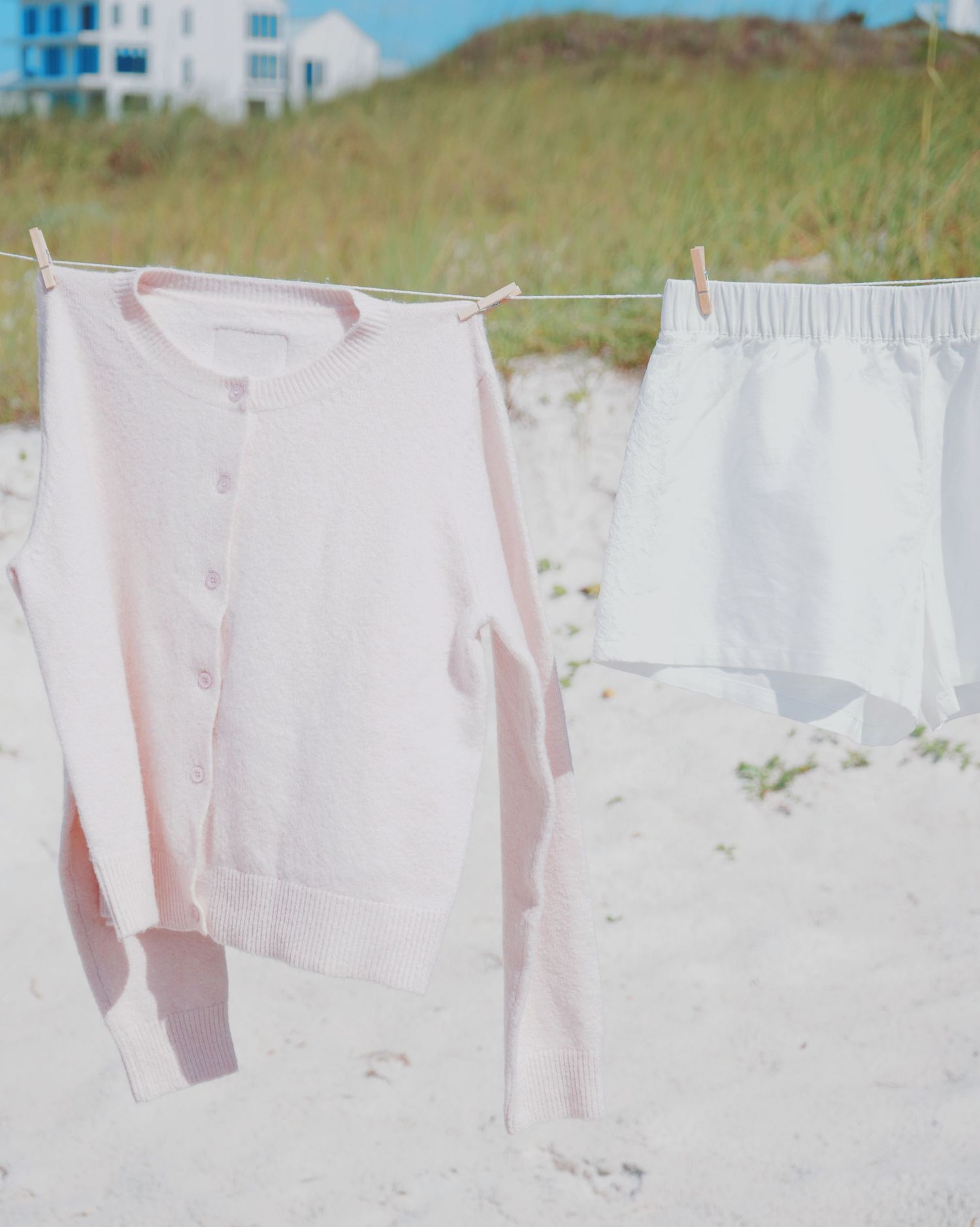 Light pink cardigan and white shorts hanging to dry on a clothesline at a sandy beach with buildings in background.