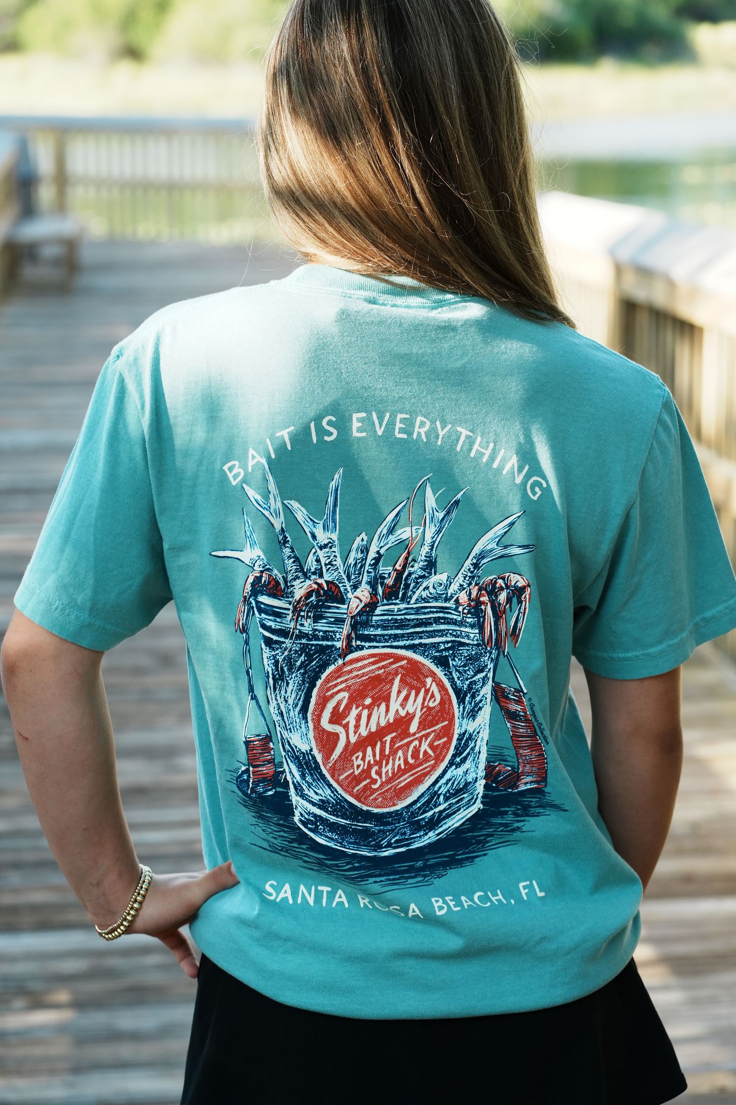 A turquoise t-shirt with 'Stinky's Fish Camp' logo and beach-themed artwork displayed on the back.