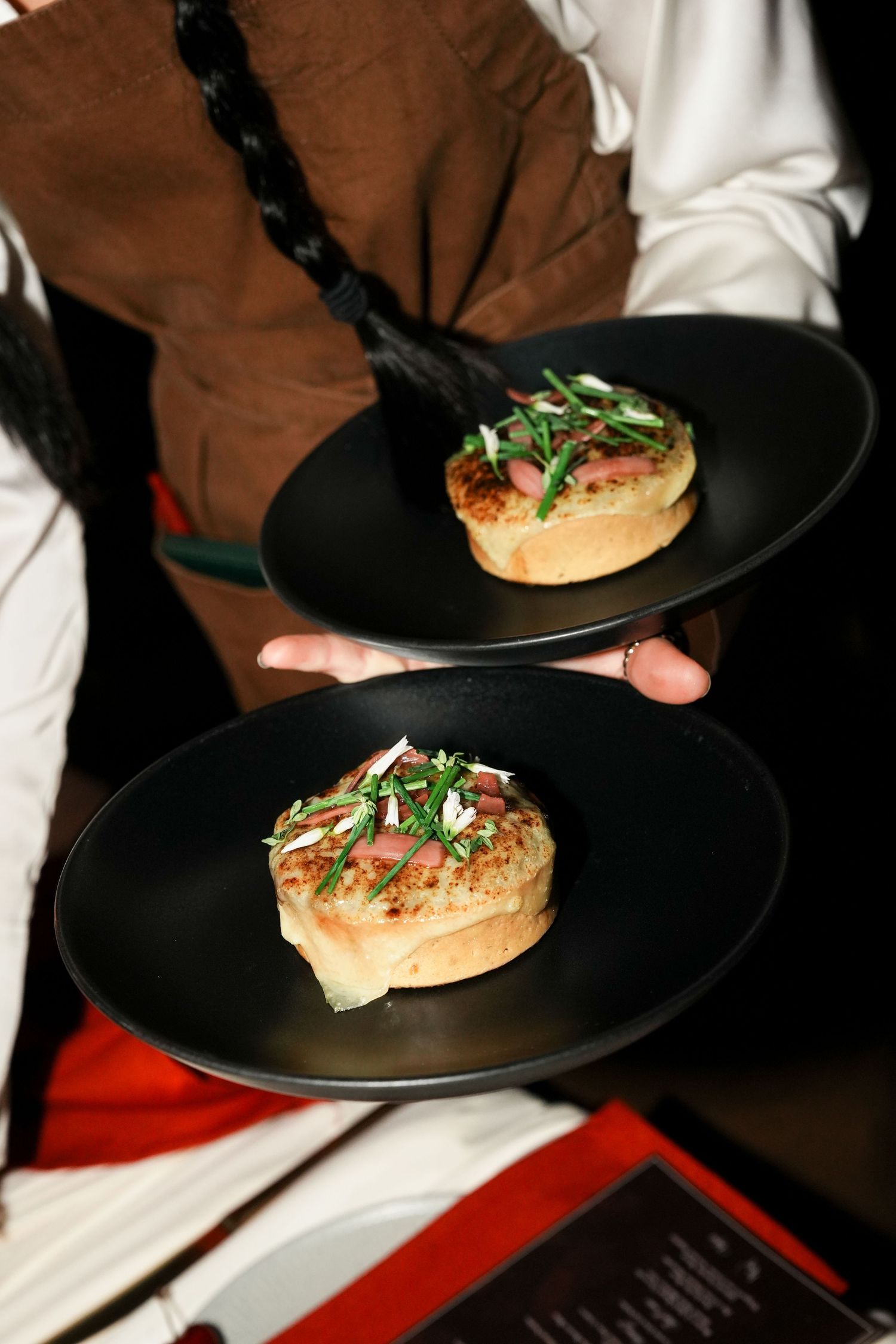 Waiter serving two black plates with gourmet open-faced sandwiches topped with fresh greens and cured meat.