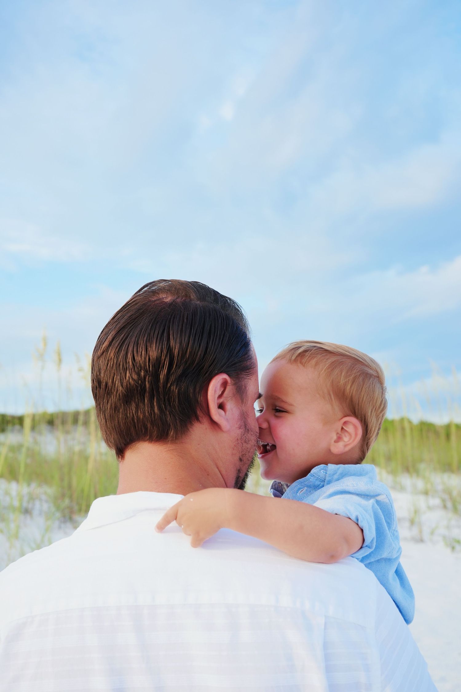 An adult and young child share tender kisses against a beachy background with white sand dunes and blue sky.