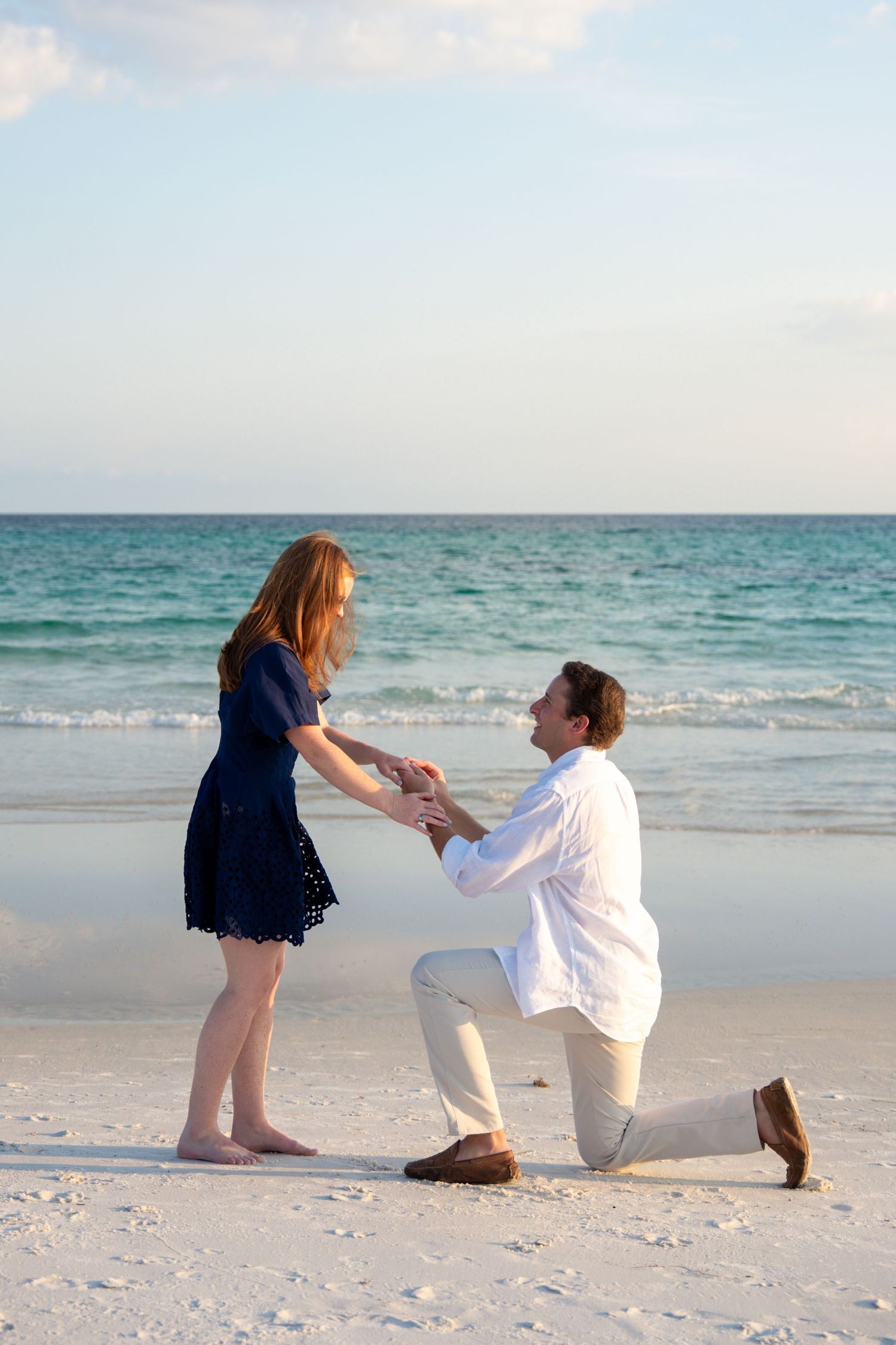 Man kneeling on one knee proposing to woman in blue dress on a white sand beach at sunset.