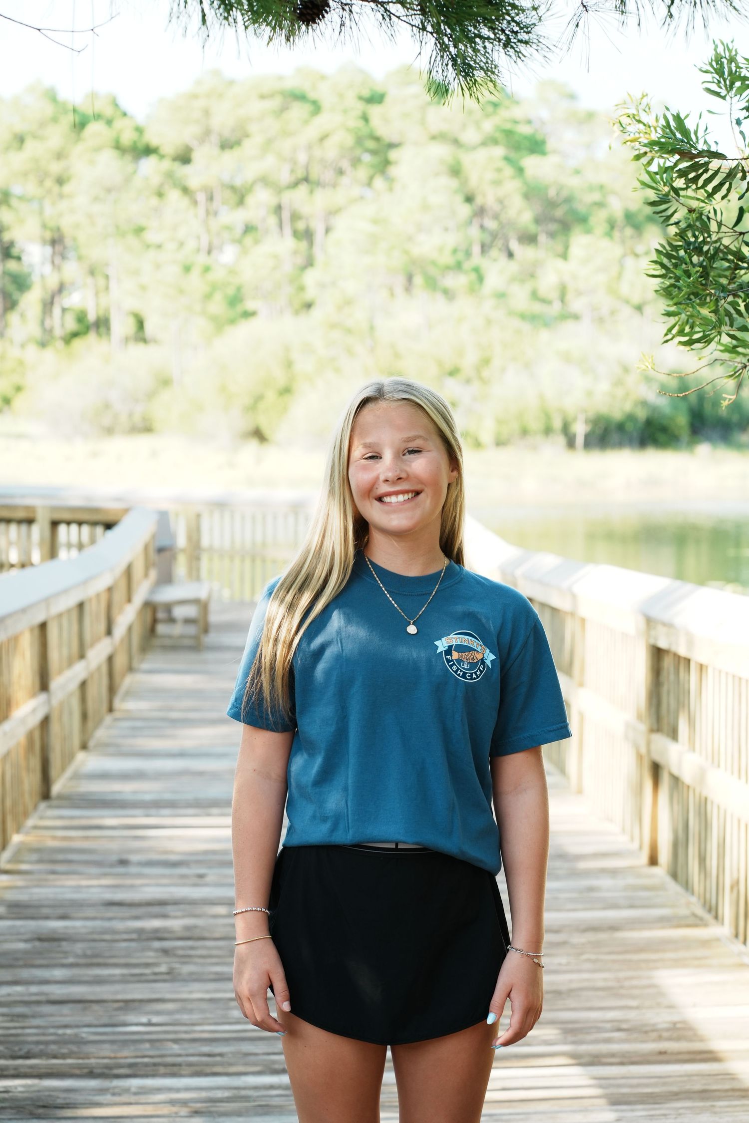 Person in navy blue shirt and black shorts standing on wooden boardwalk with trees in background.