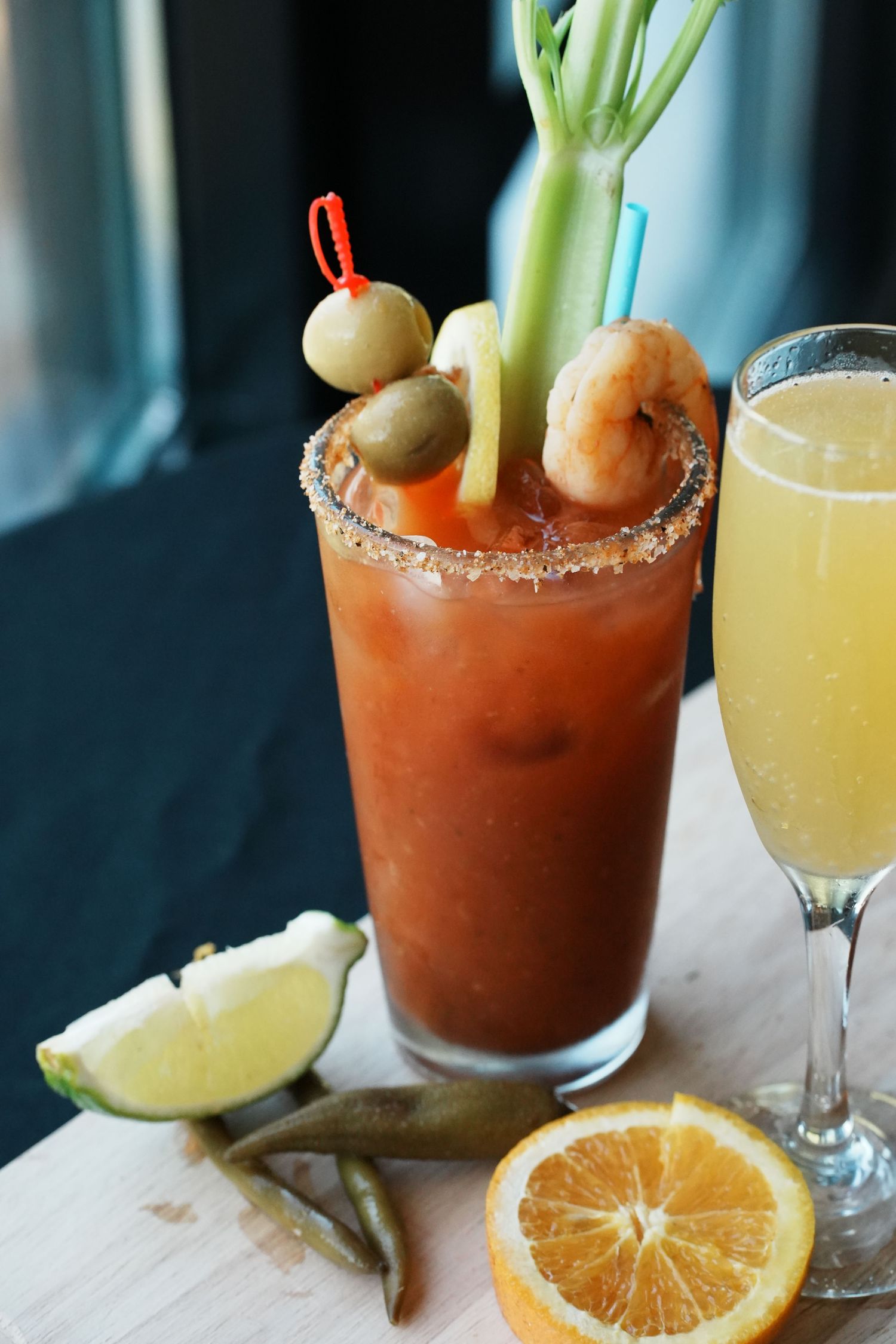 Bloody Mary cocktail garnished with celery, olives and shrimp alongside a mimosa on a wooden surface.