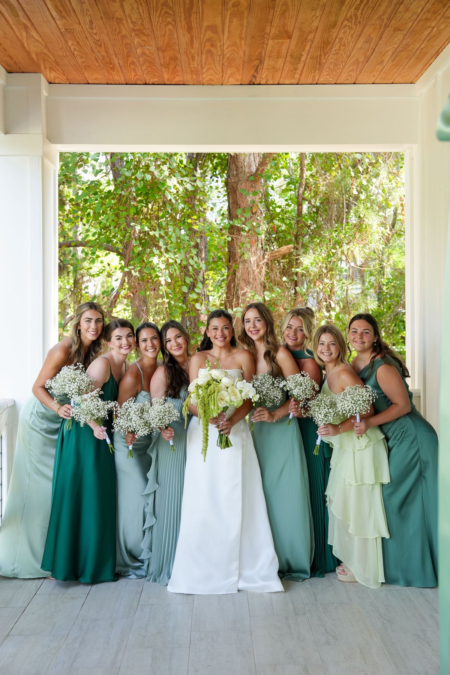 Bride in white gown poses with eight bridesmaids in mismatched green dresses holding baby's breath bouquets outdoors.
