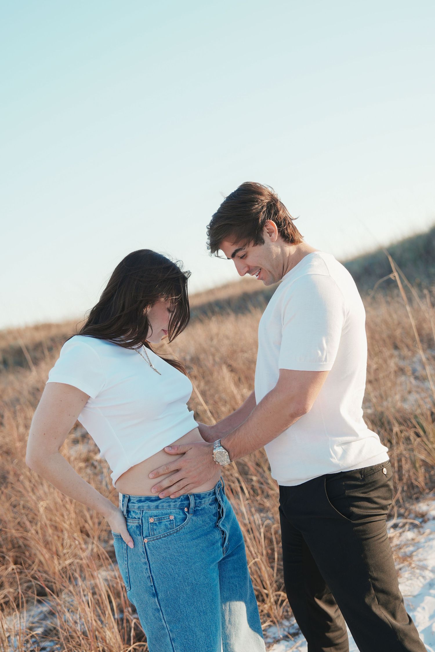 A couple in white shirts and jeans share tender moments in a sunlit field of tall grass by the water.