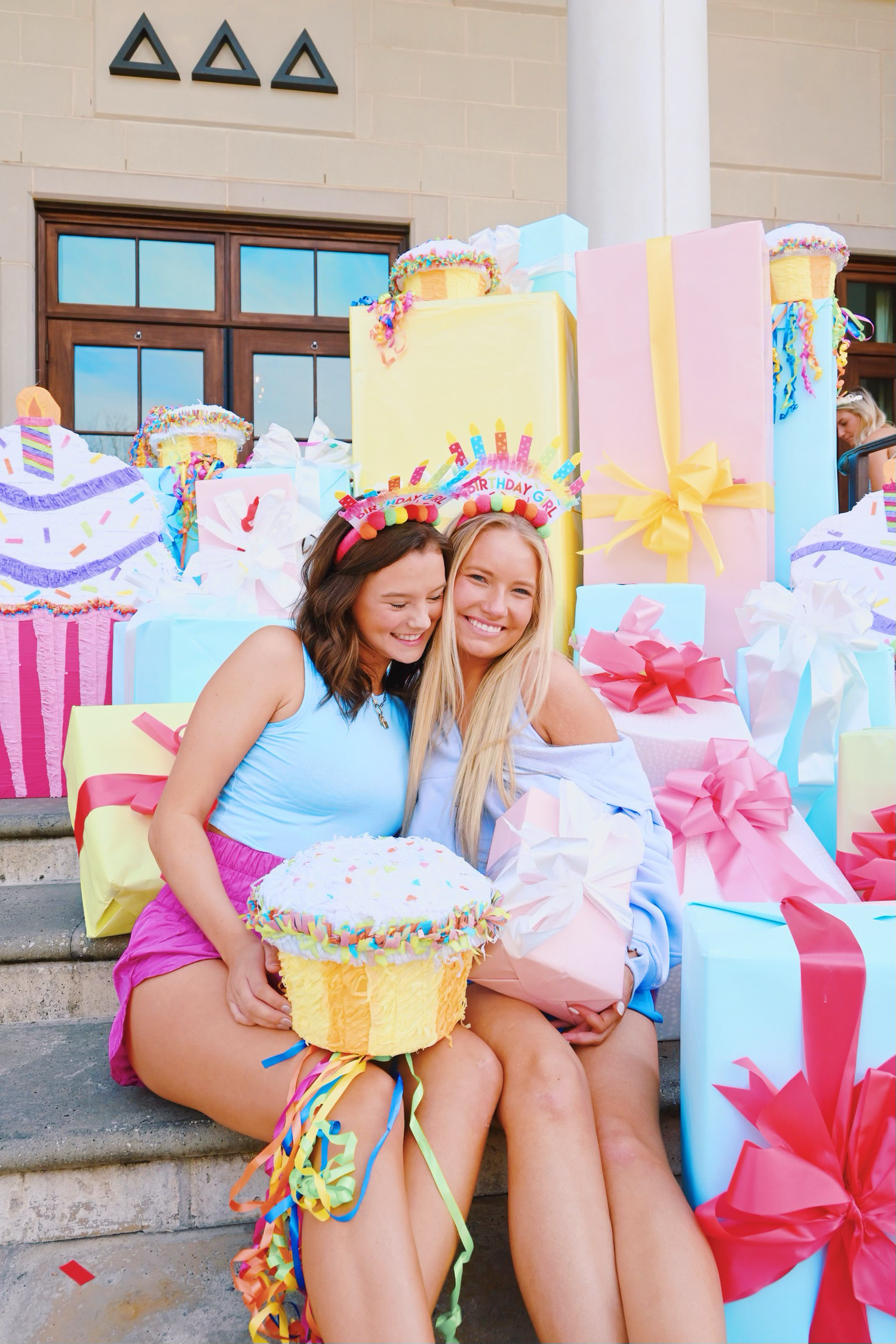 Two cheerleaders sitting on steps surrounded by colorful gift boxes during a celebration photoshoot.