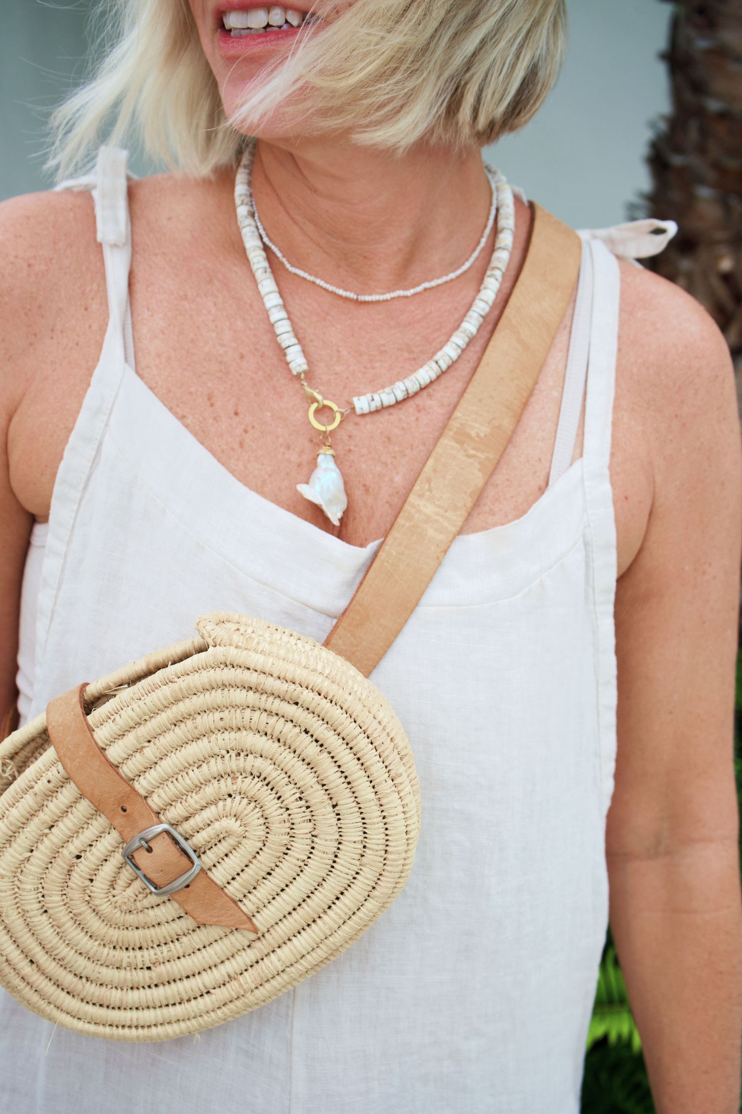 Close-up of white tank top with layered shell necklaces and oval straw crossbody bag with leather strap.