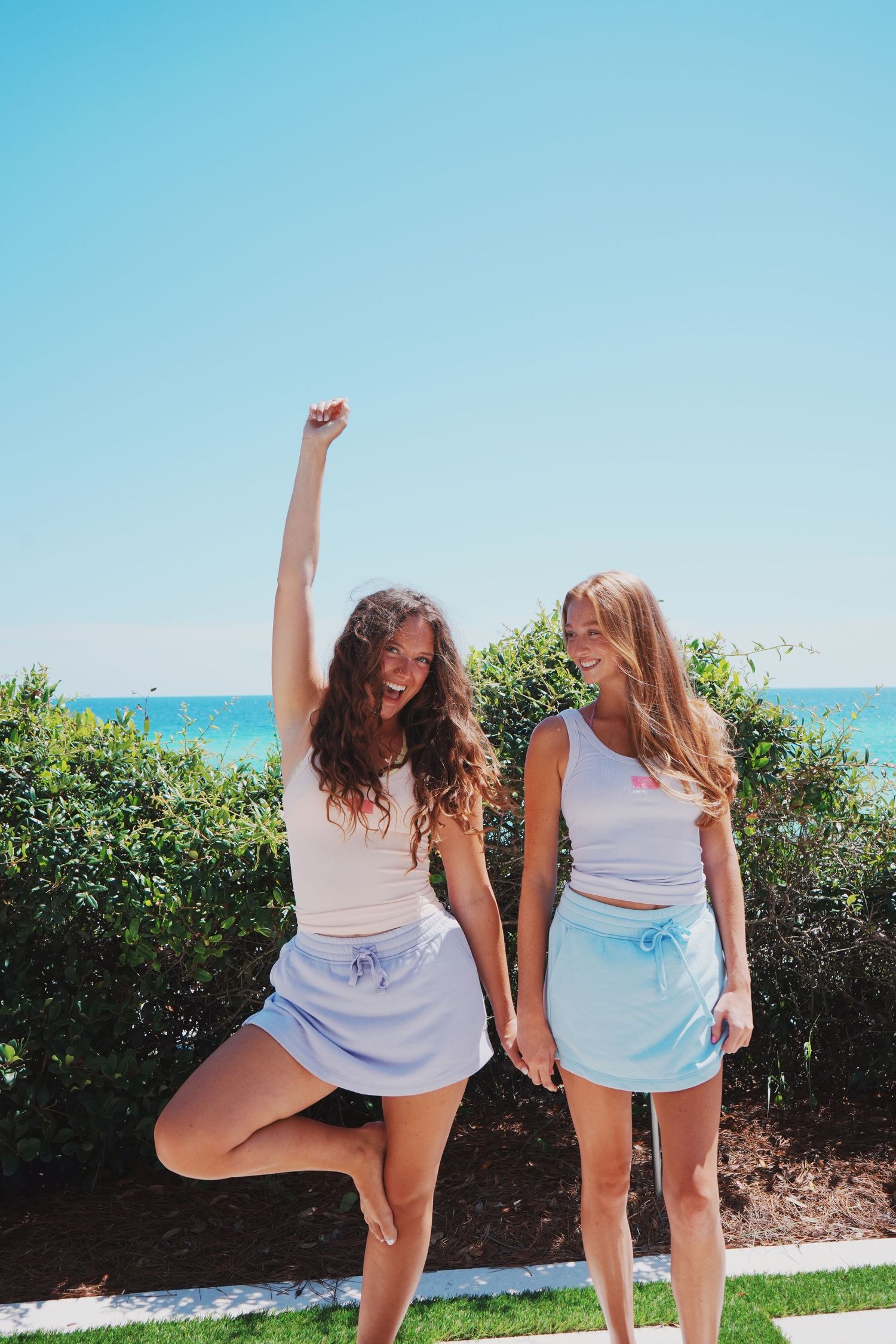 Two friends in white outfits strike playful poses outdoors against a backdrop of blue sky and greenery.