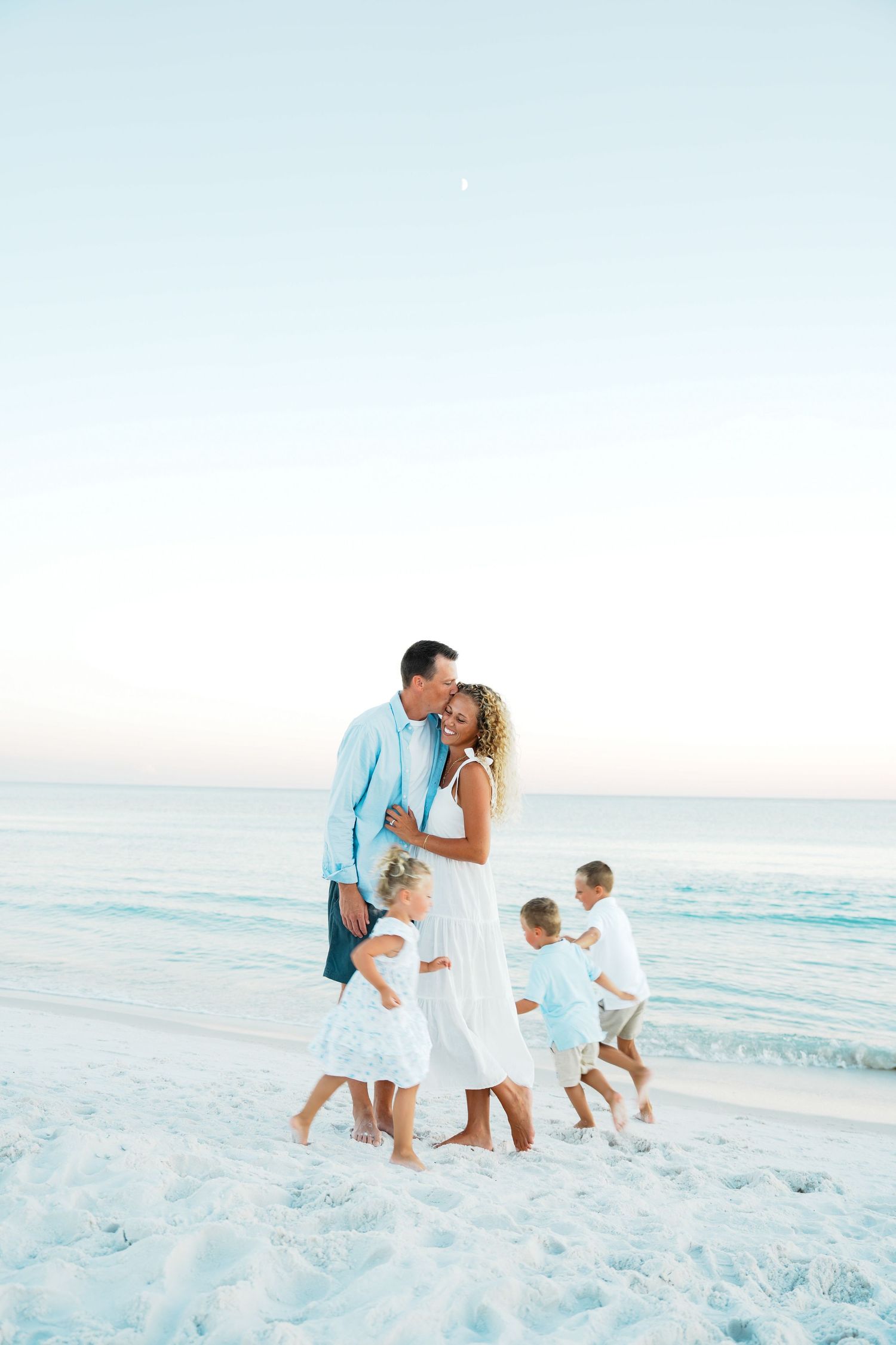 Family in matching white and blue outfits walking together on a pristine white sand beach at sunset.