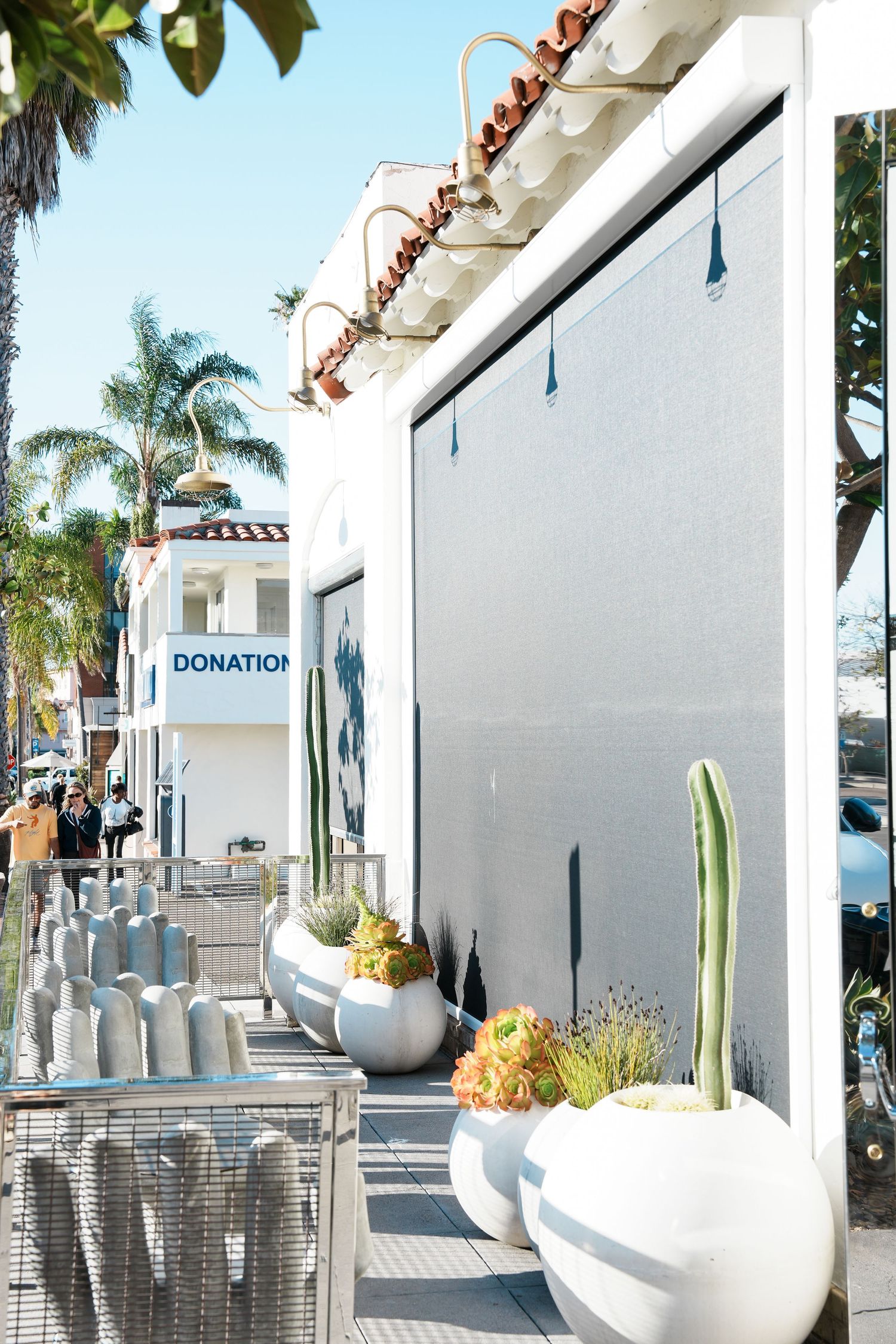 White modern storefront with large planters featuring cacti and succulents, beside a donation center with palm trees.
