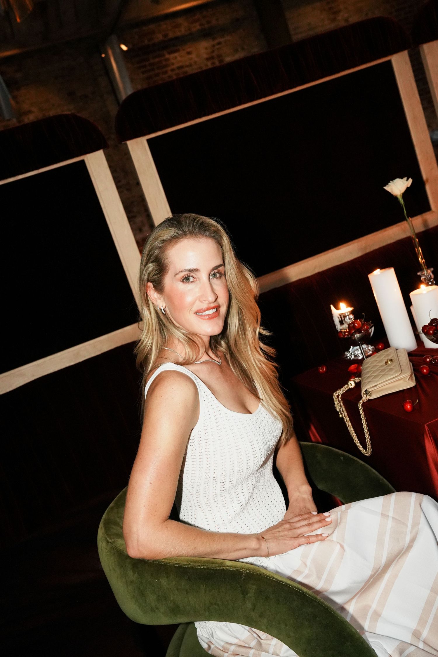 Elegant blonde woman in white dress sitting in green velvet chair at candlelit dinner event.