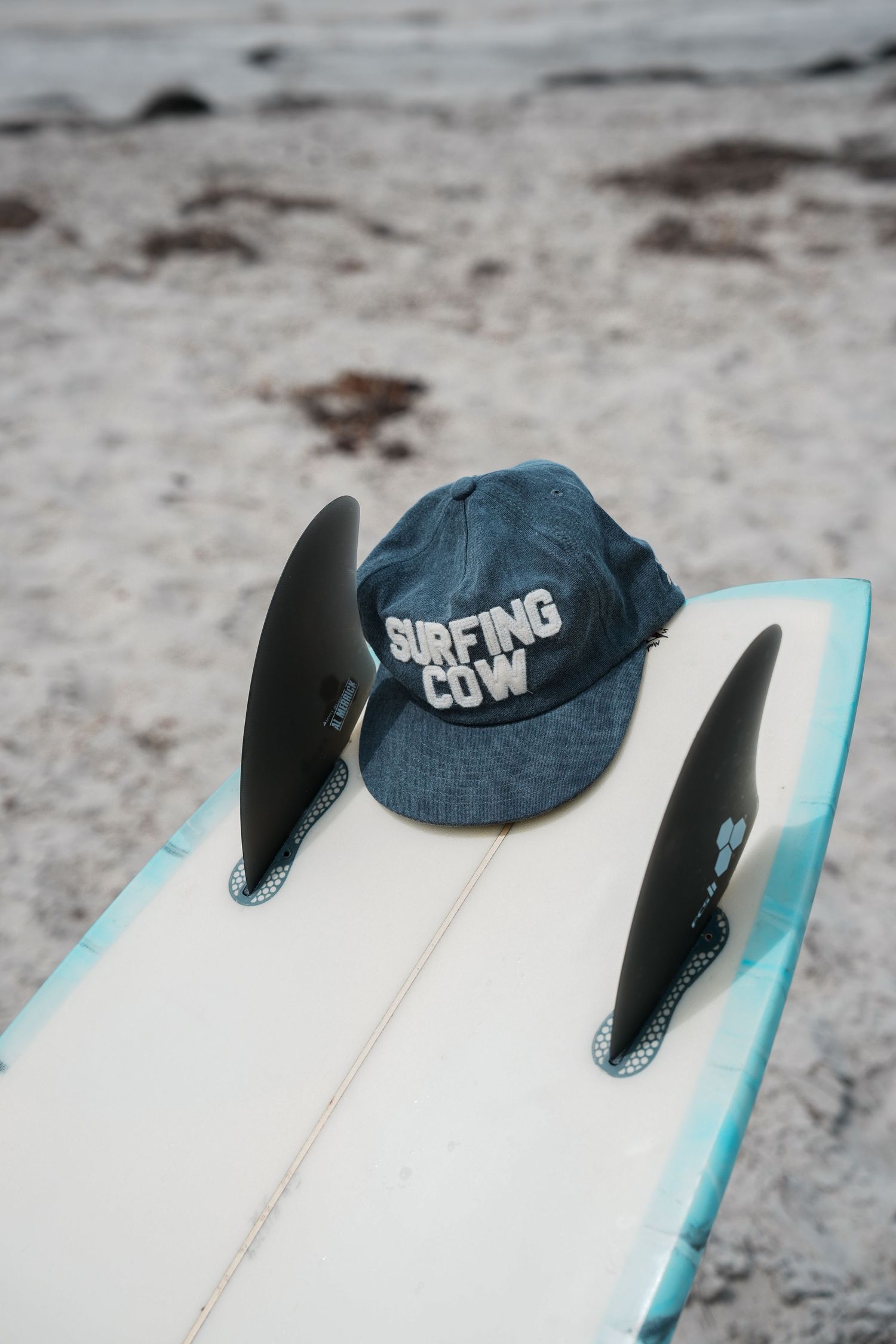 A denim Surfing Cow cap resting on a white surfboard with black fins on a sandy beach.