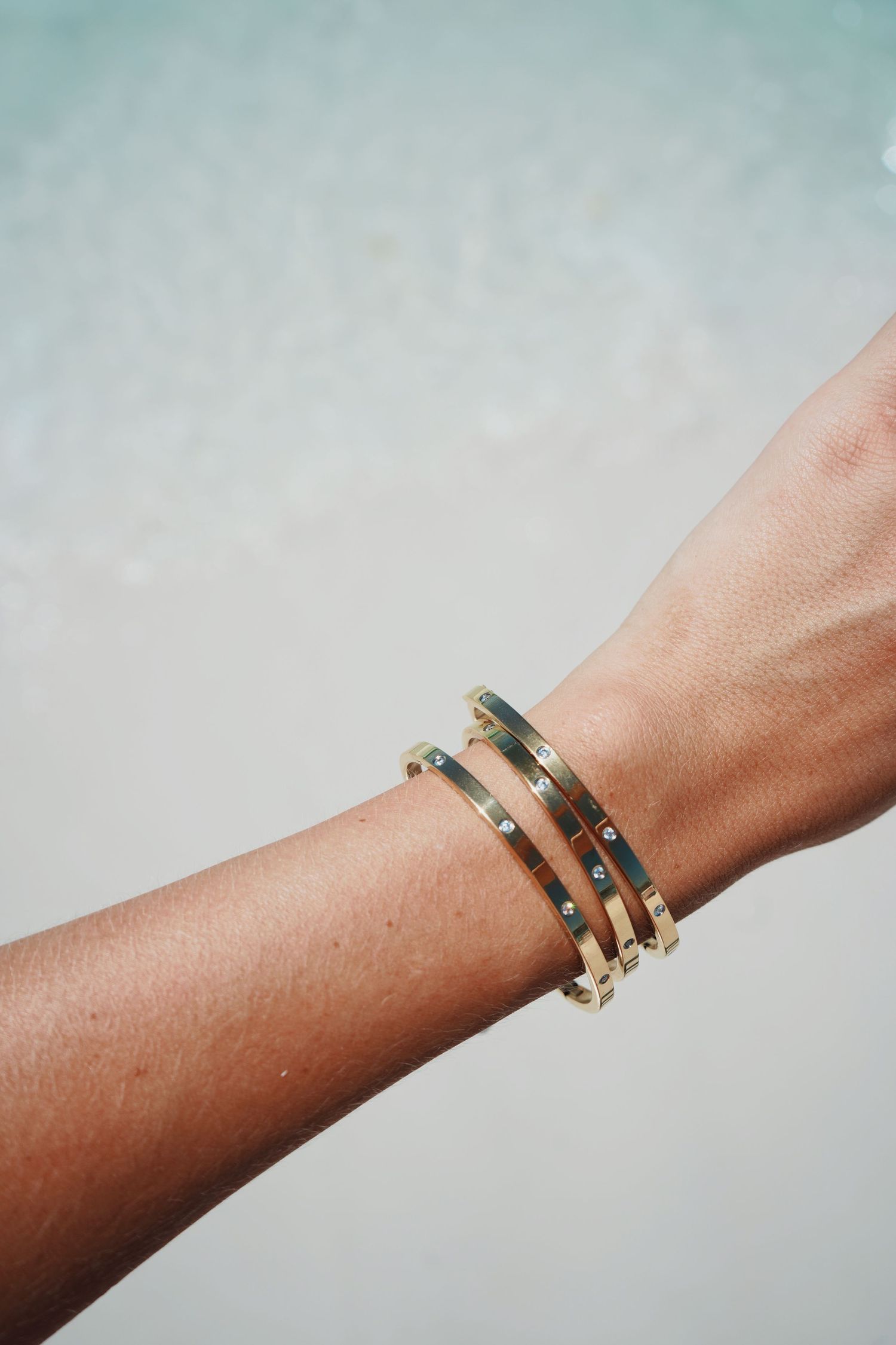 A series of delicate gold and gemstone stacking bracelets shown on a wrist against a light blue background.