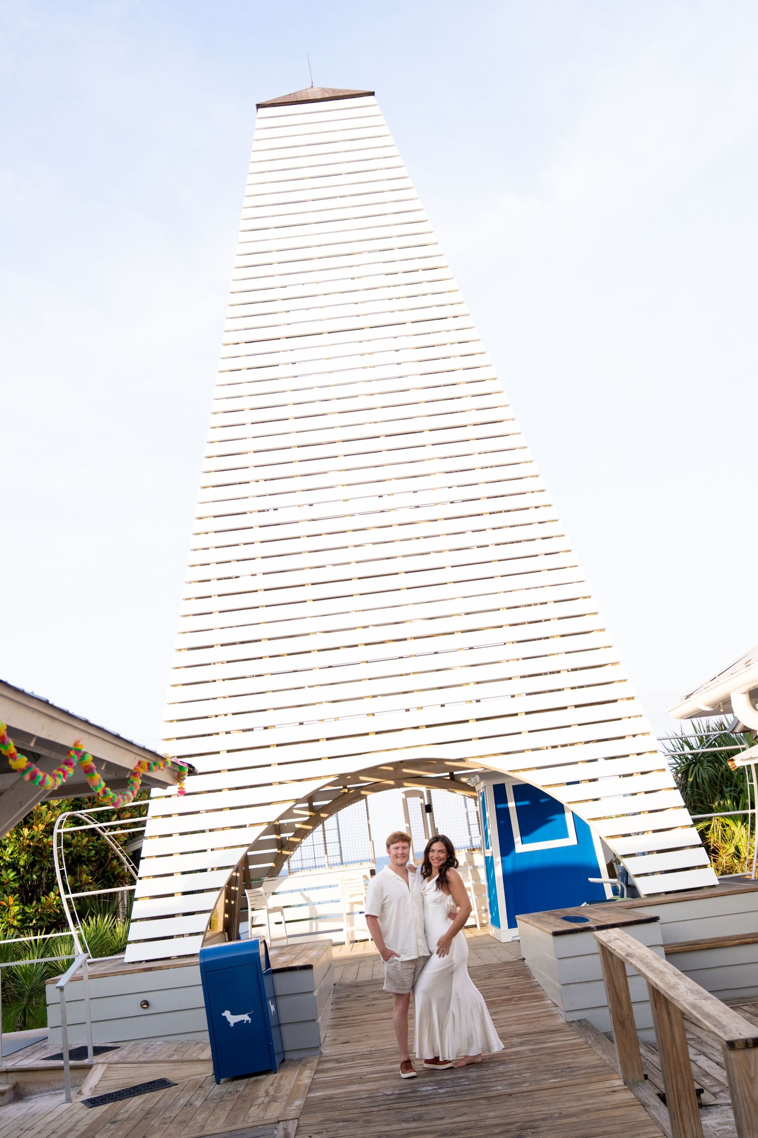 Couple in white attire posing on wooden boardwalk before large lattice pyramid structure at coastal venue.