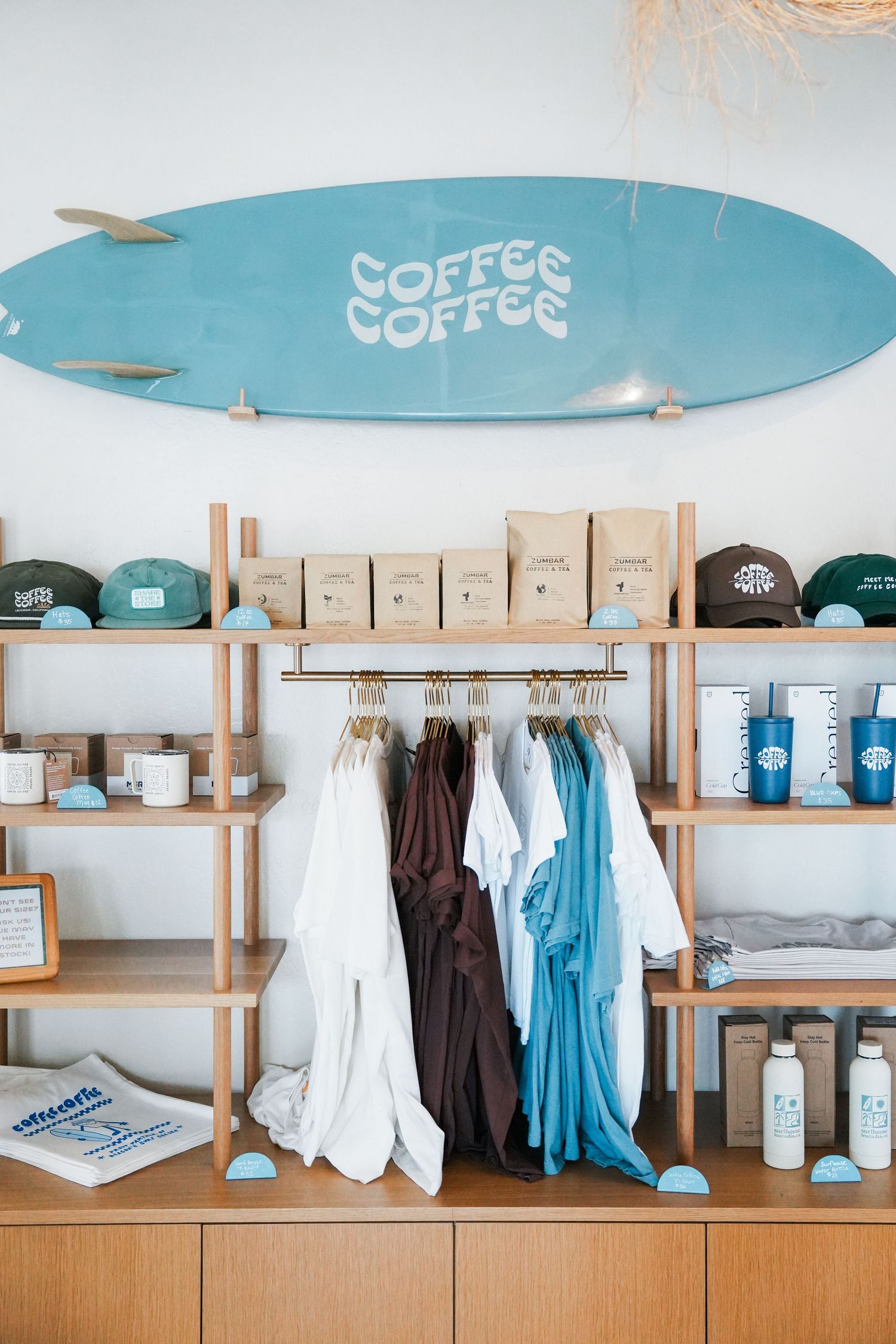 A surf shop interior with a blue surfboard, clothing racks, and coffee merchandise on wooden shelving.