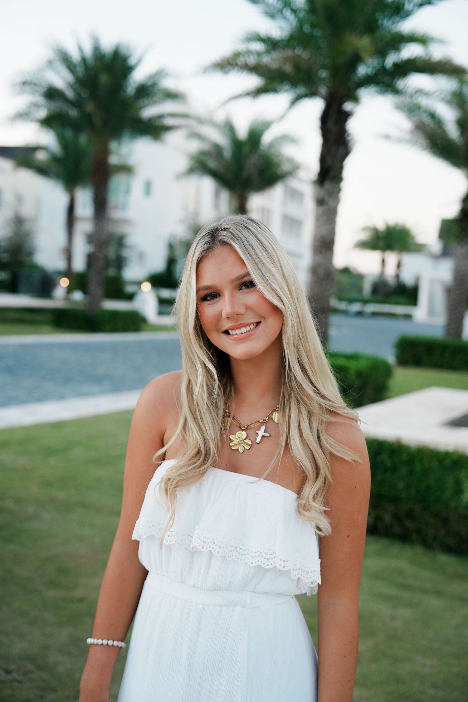 Woman in white summer dress poses outdoors with palm trees and white buildings in background.