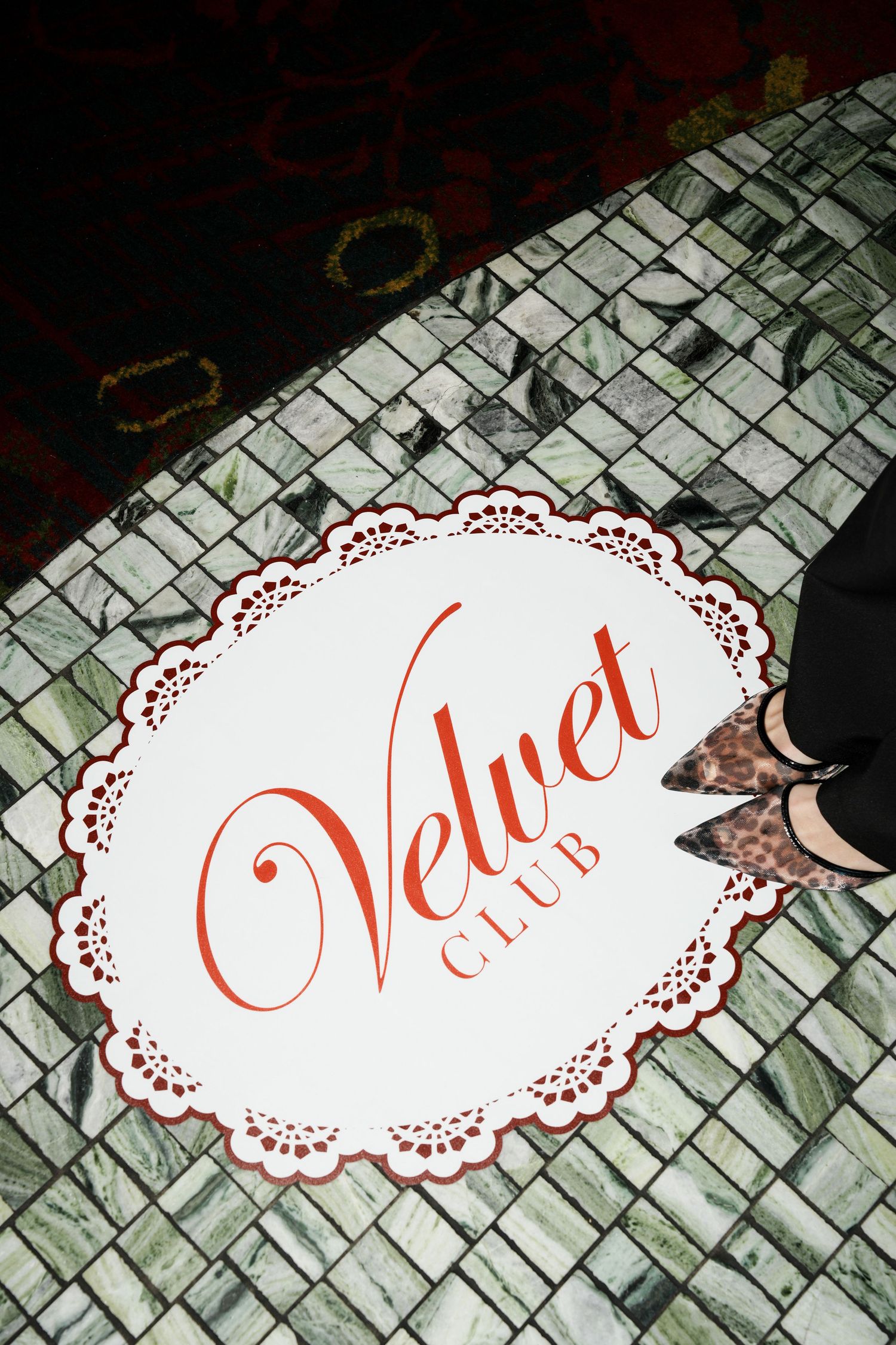 Velvet Club logo on decorative tile floor with lace border design, partial view of leopard print heels.