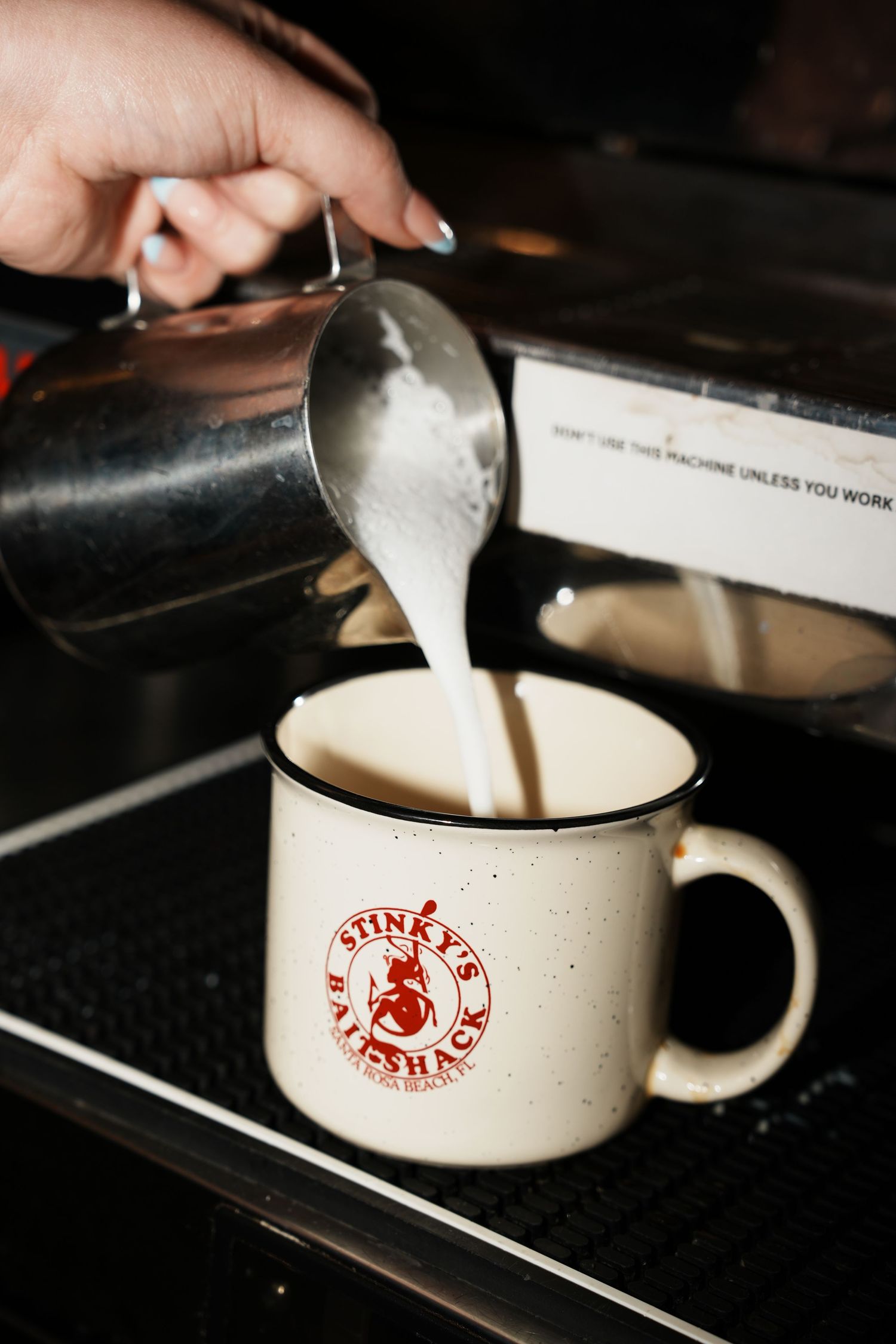 Pouring steamed milk into a white coffee cup with red logo at a cafe counter.