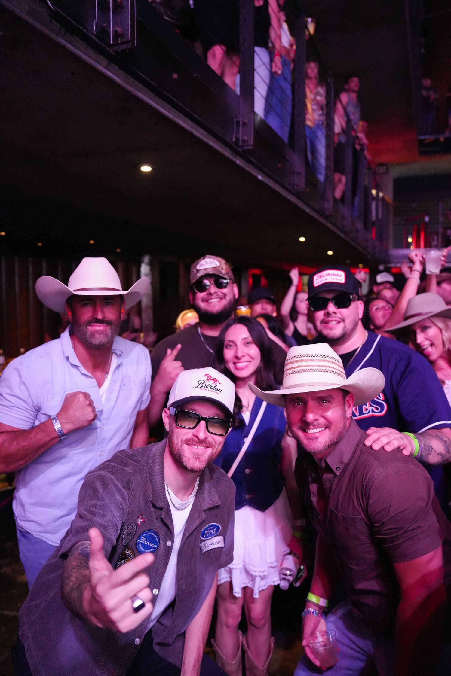A group of concert attendees in cowboy hats and sunglasses smile and pose together at a music venue.