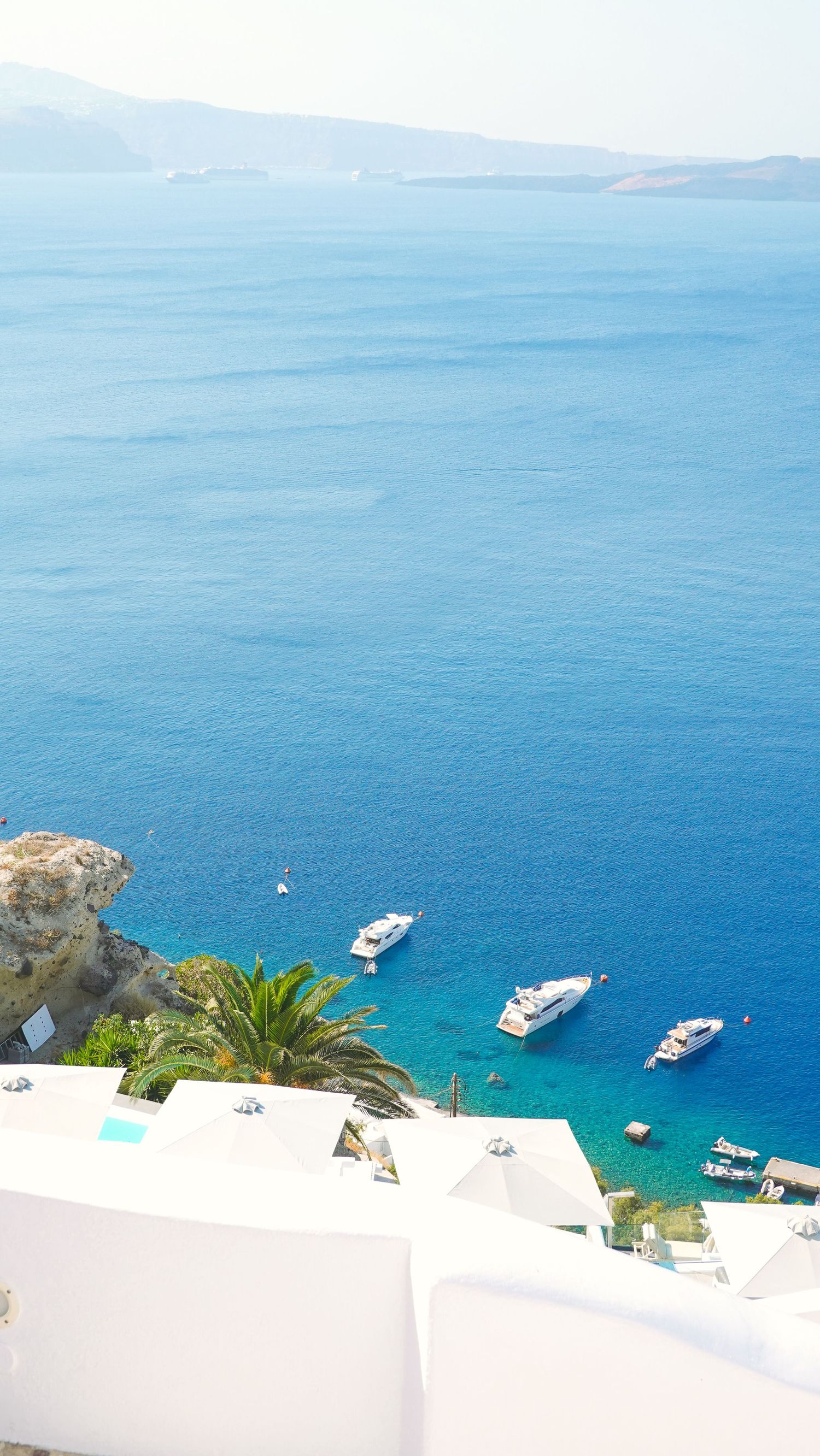 Luxury yachts anchored in crystal clear Mediterranean waters below white buildings on a coastal cliff.