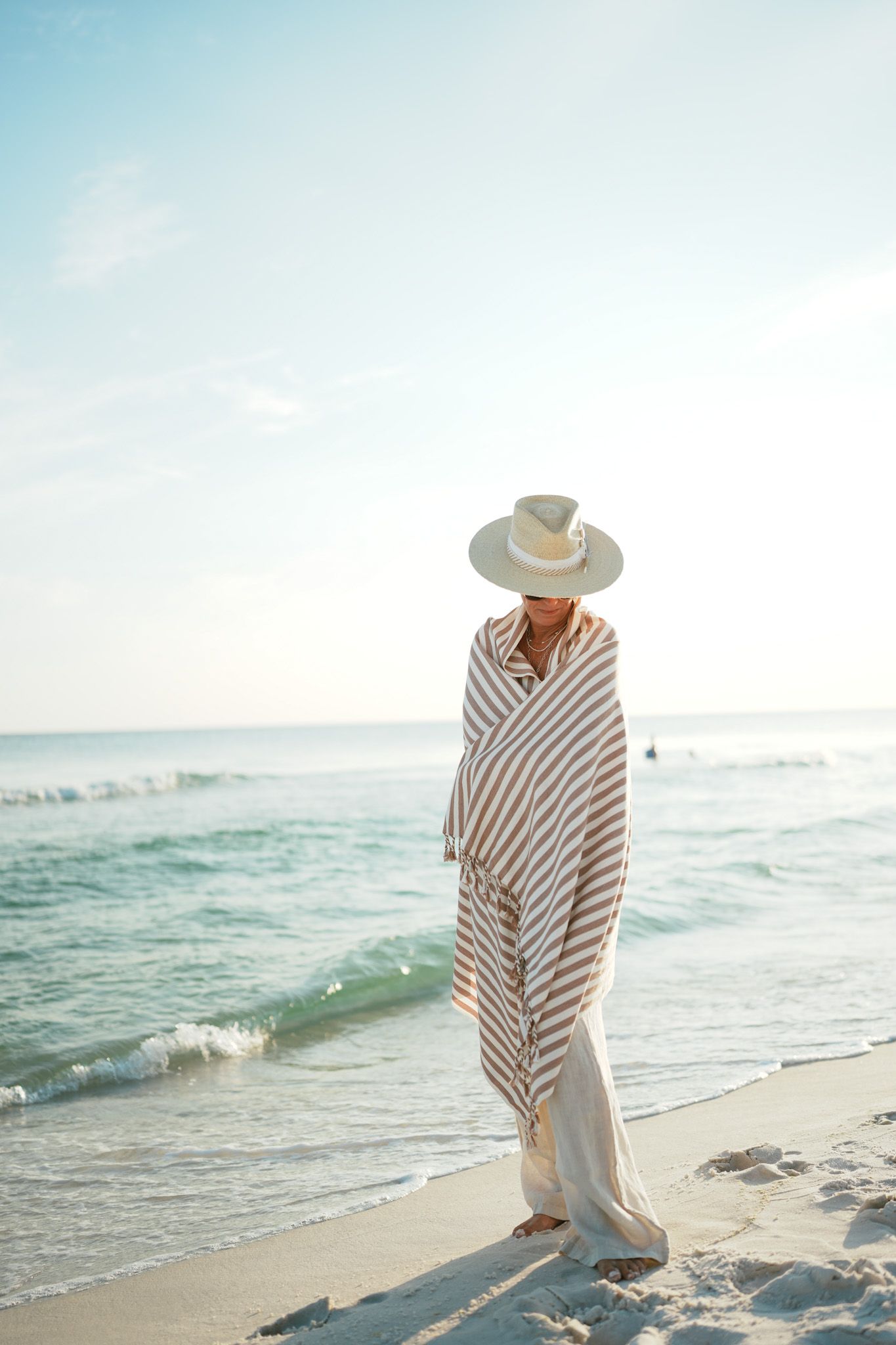 A person in a striped poncho and sun hat walks along a serene beach at sunset with gentle waves rolling in.