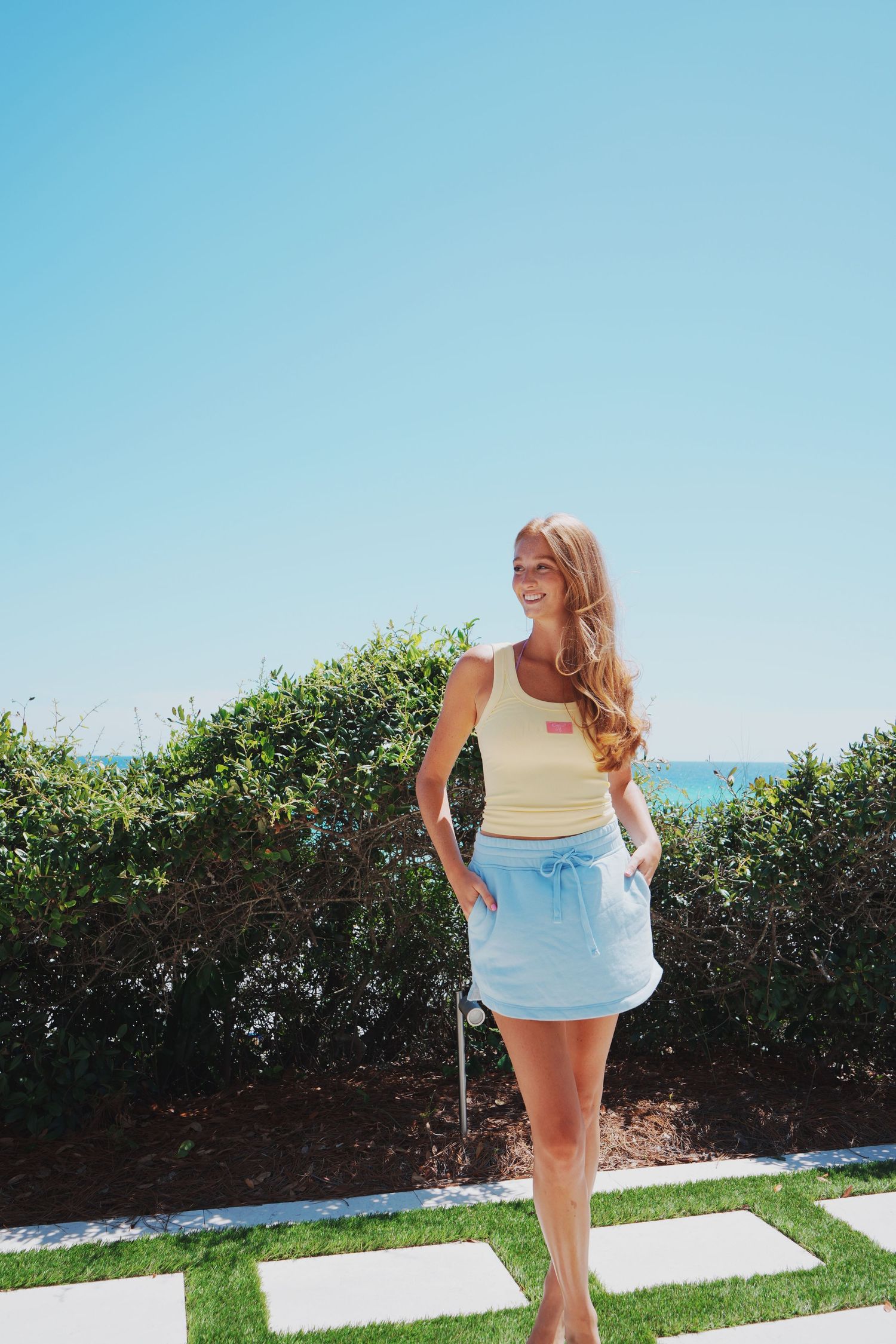 Person in casual summer outfit standing on paved path with ocean view in background.