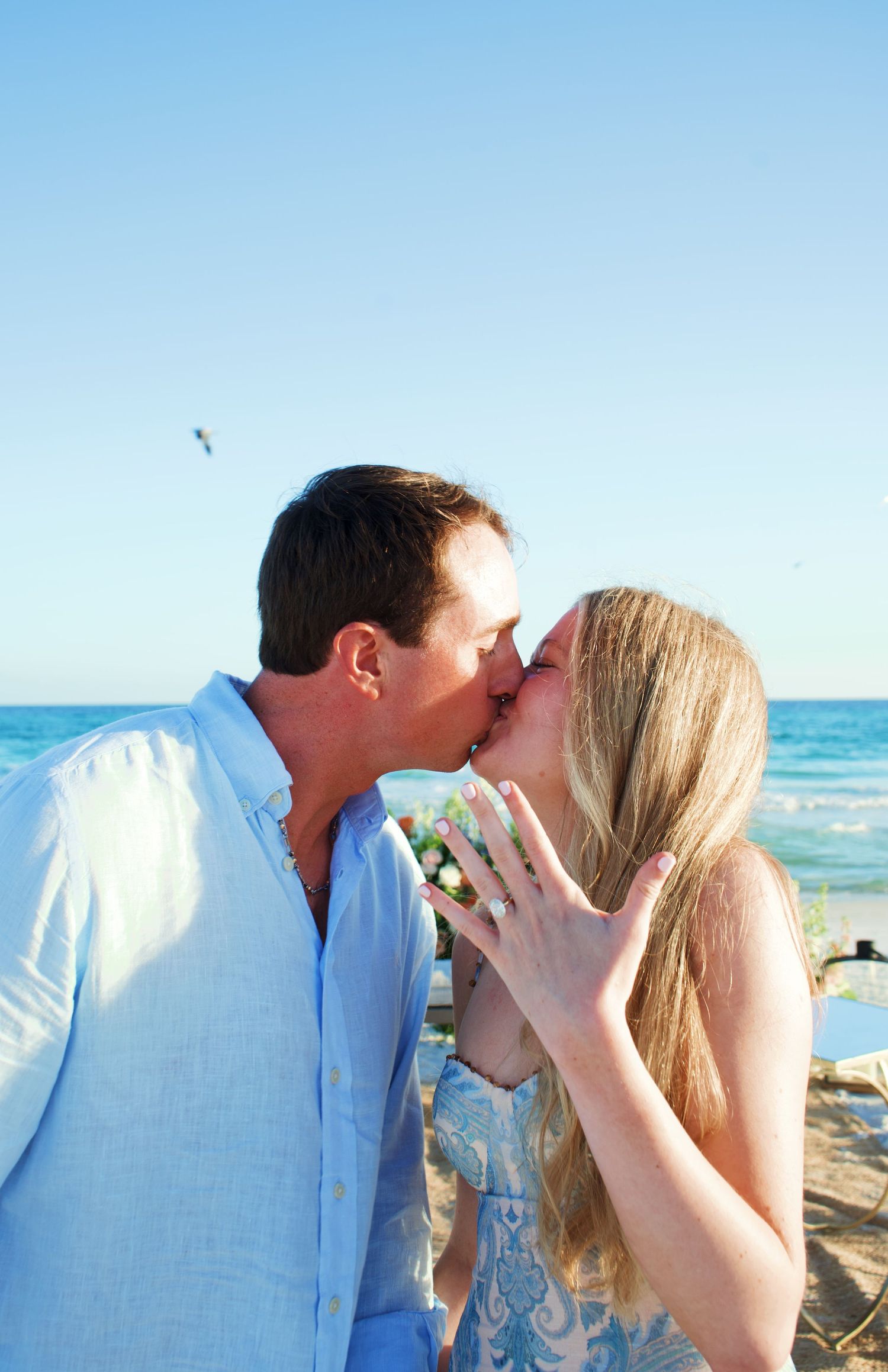 Romantic couple shares intimate kiss on sunny beach with ocean waves and blue sky in background.