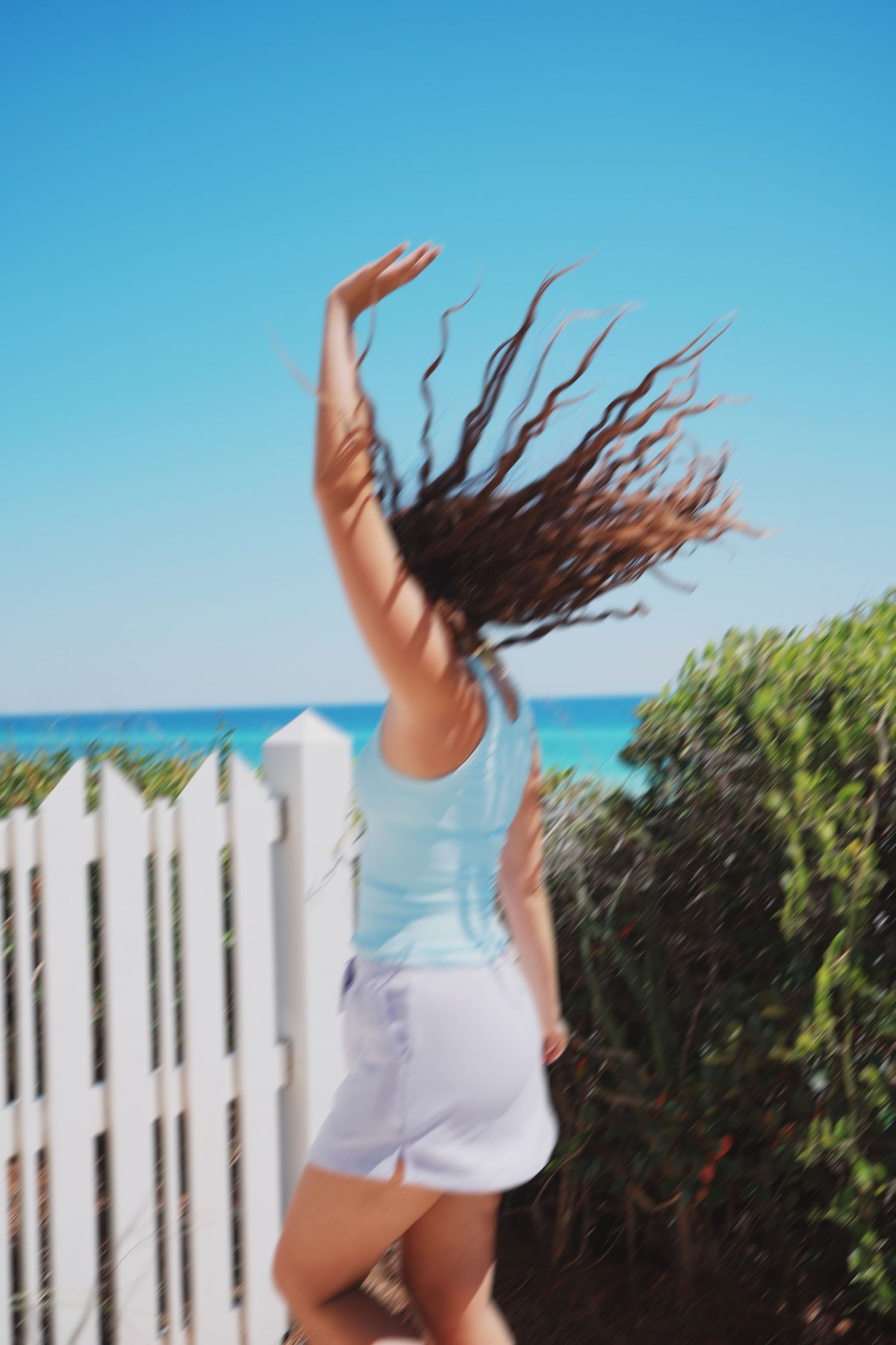 Person with flowing hair jumps joyfully by white fence with ocean view.