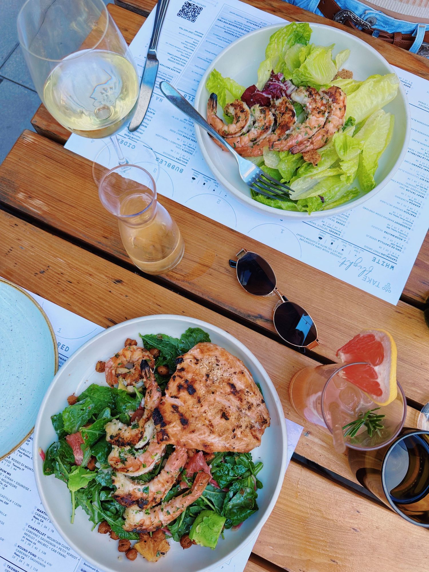 Fresh salads with grilled protein and glasses of wine on wooden table with menu and sunglasses.