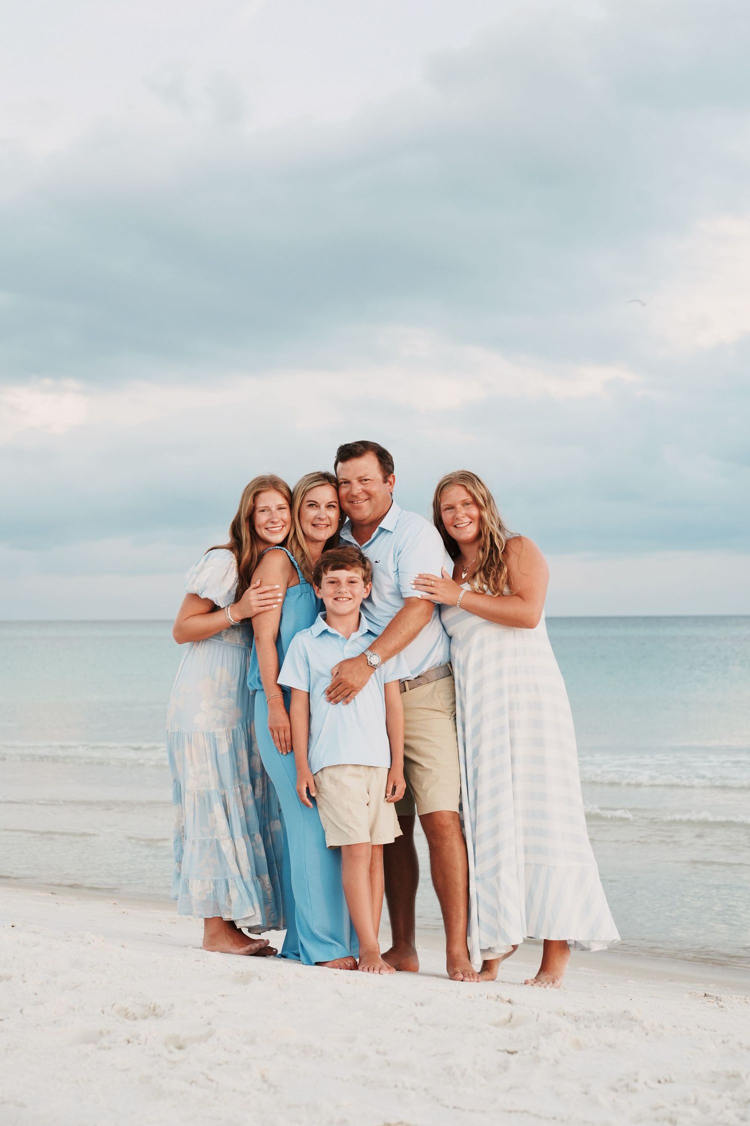 Family group wearing light blue and white clothing poses together on sunny beach with ocean backdrop.