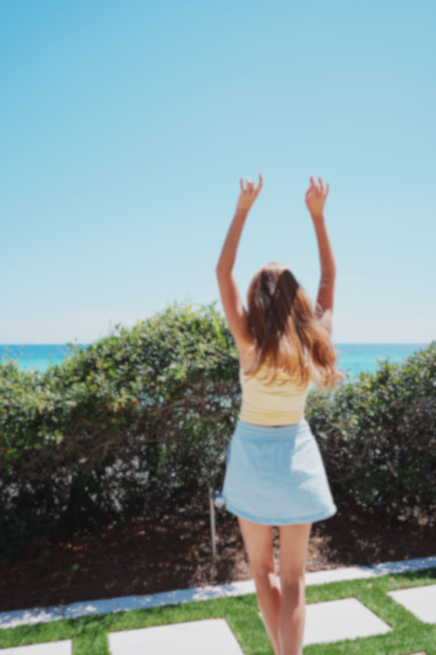 Person in yellow top and light blue skirt raises arms joyfully by the ocean with bushes in background.